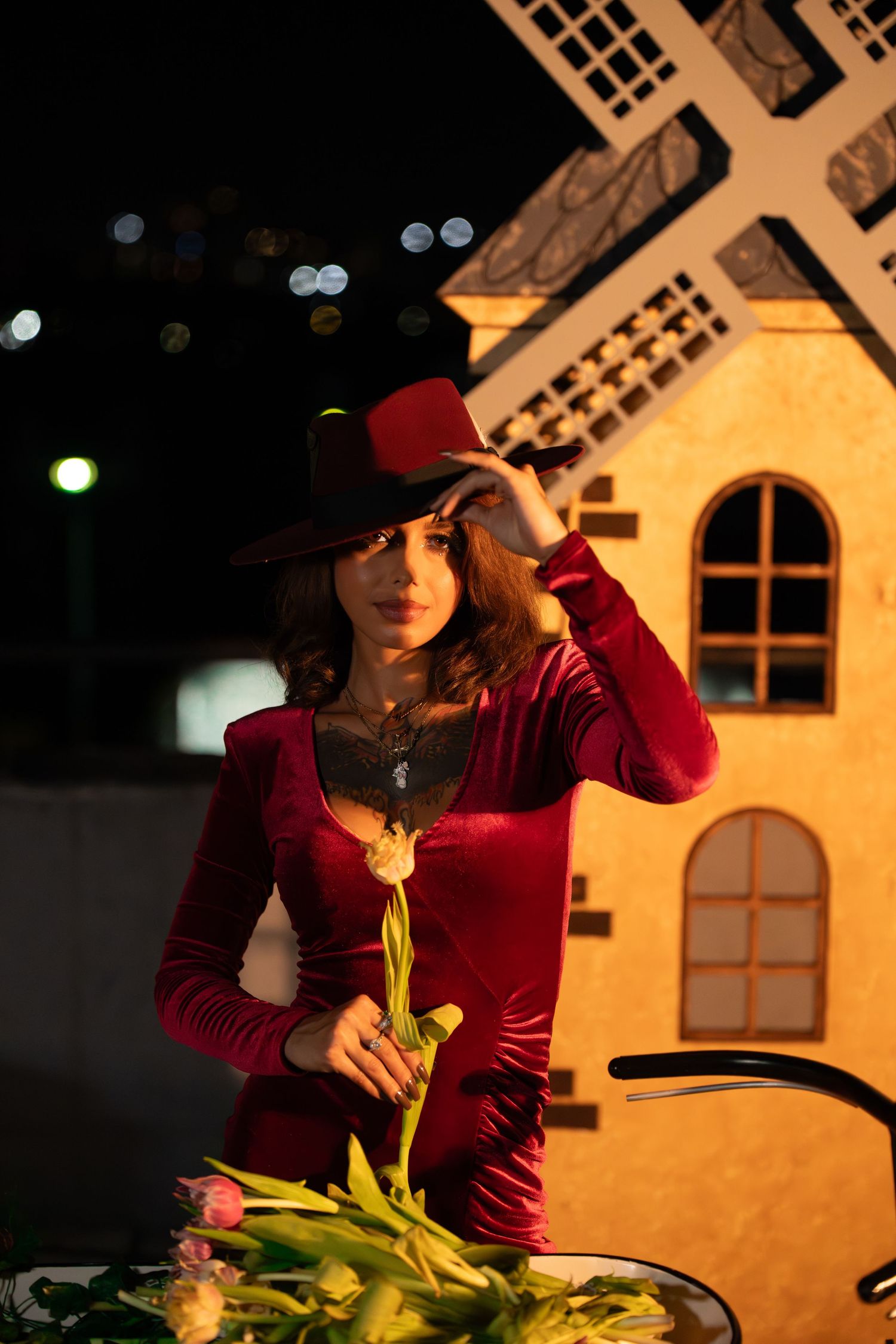 Figure in red dress and hat stands near a lit windmill building in evening light.