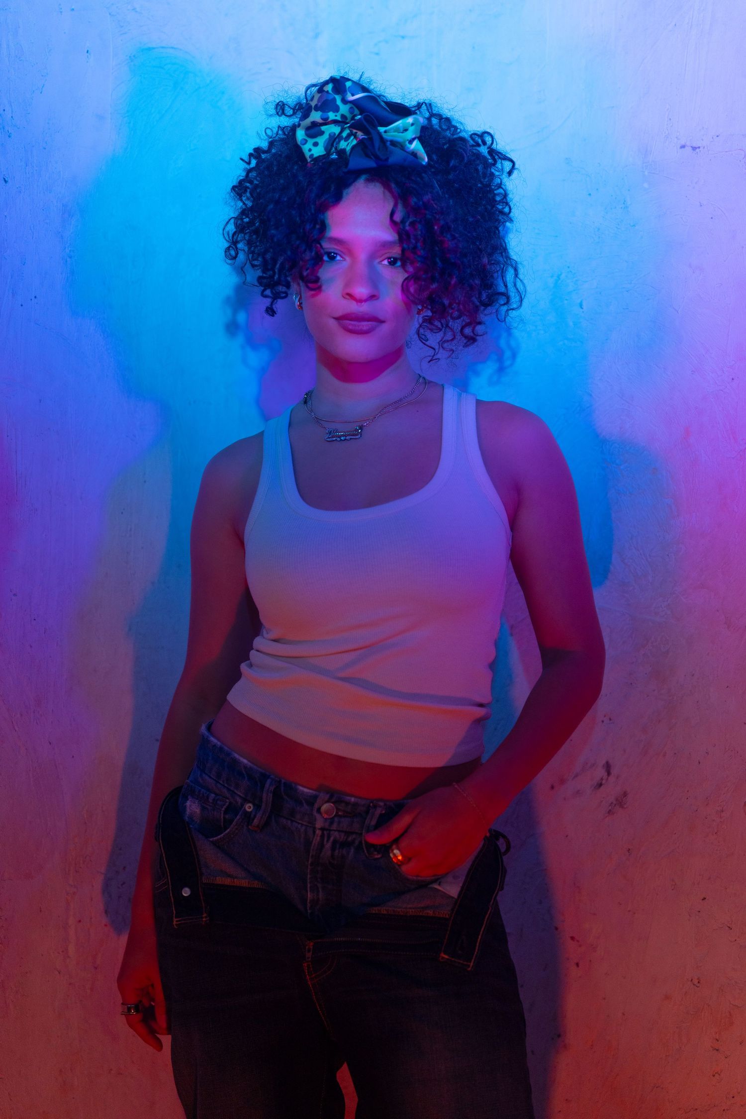 A person with curly hair poses in a white tank top against a blue and red gradient background in a photo series.