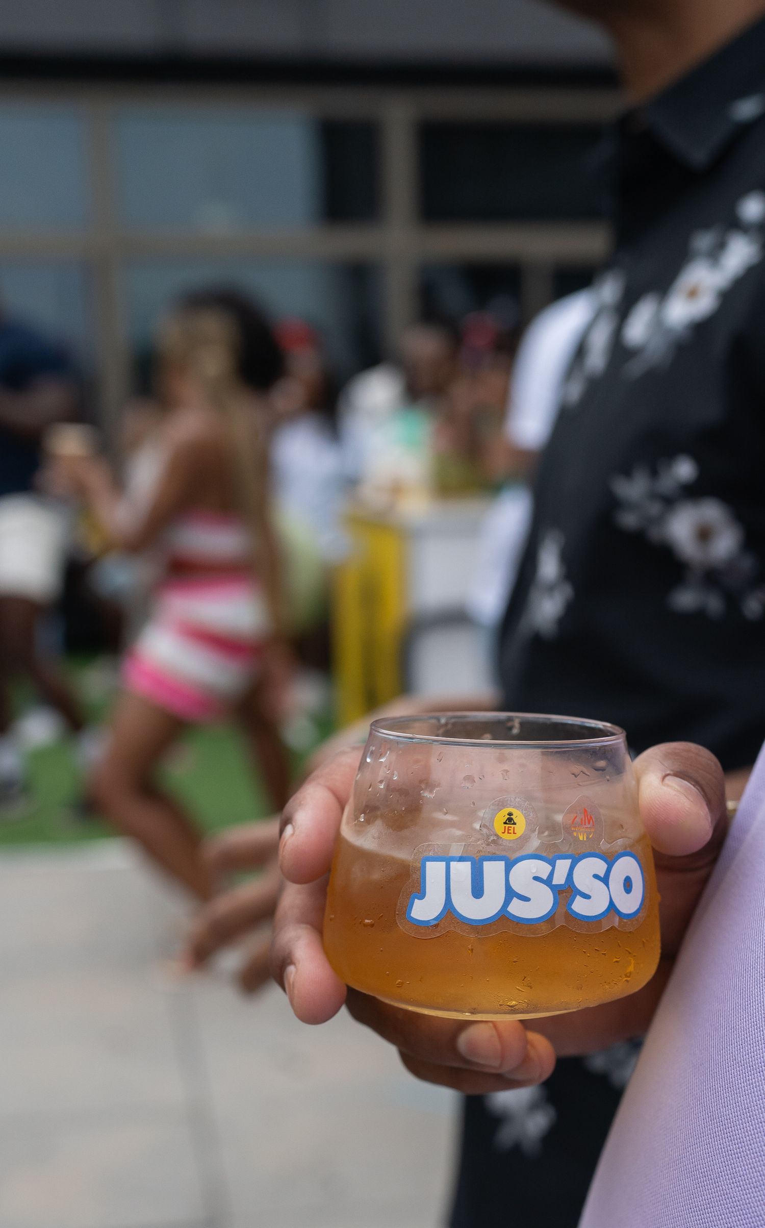Close-up shot of a hand holding a glass of Jus'So branded beverage at an outdoor event.