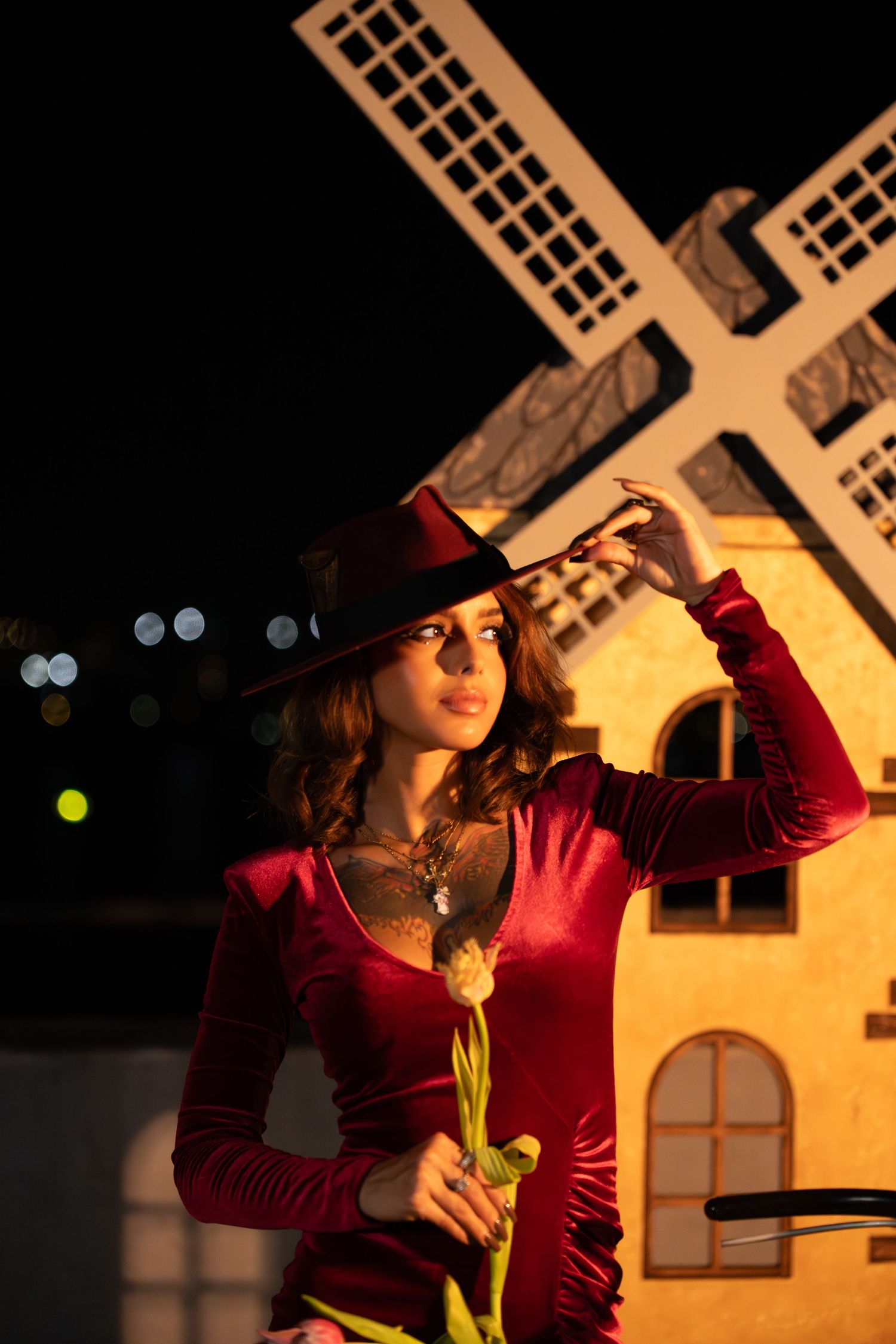 Woman in red velvet dress and black hat poses dramatically against illuminated windmill structure at night.