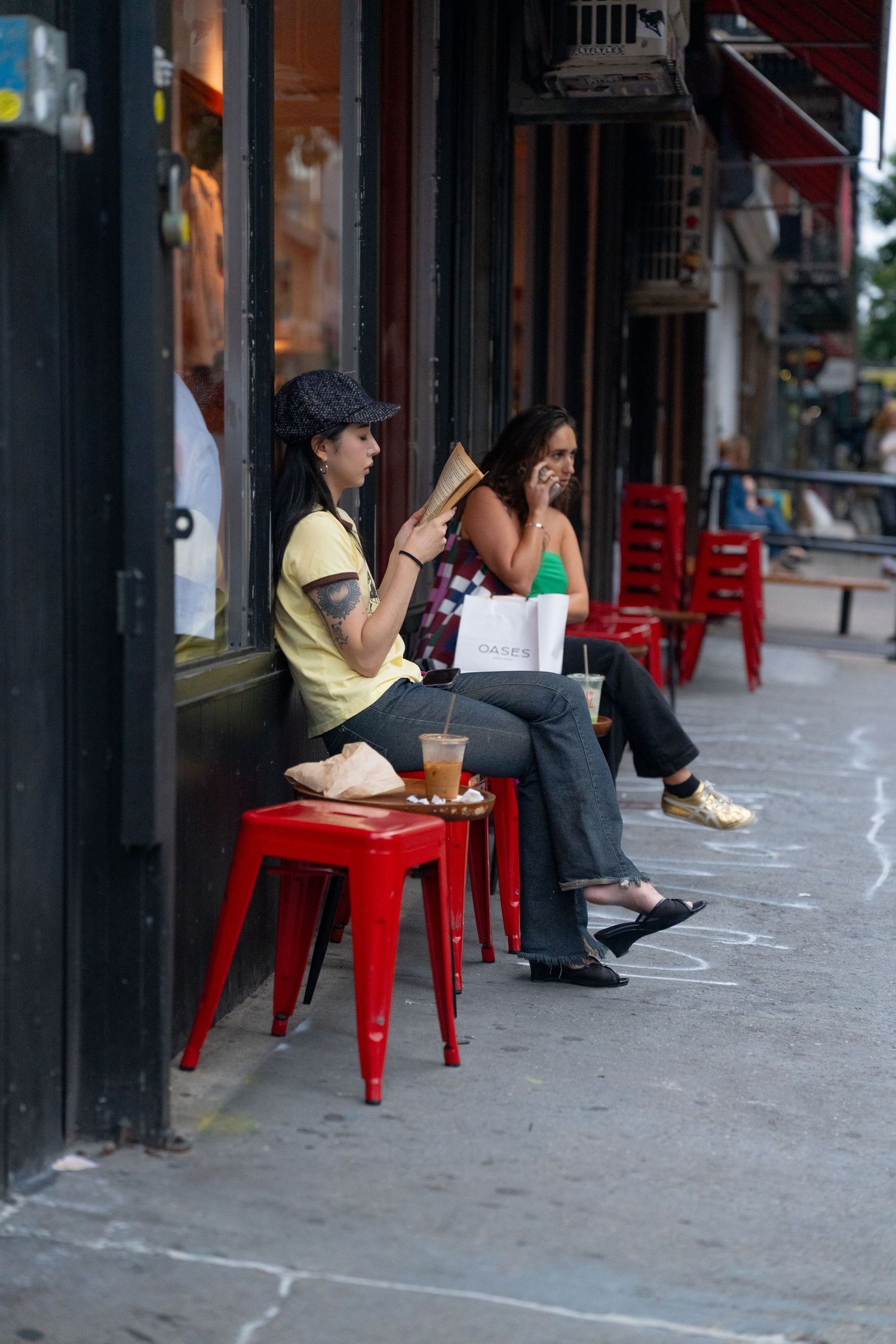 People relaxing on red metal chairs outside a storefront on a city sidewalk.
