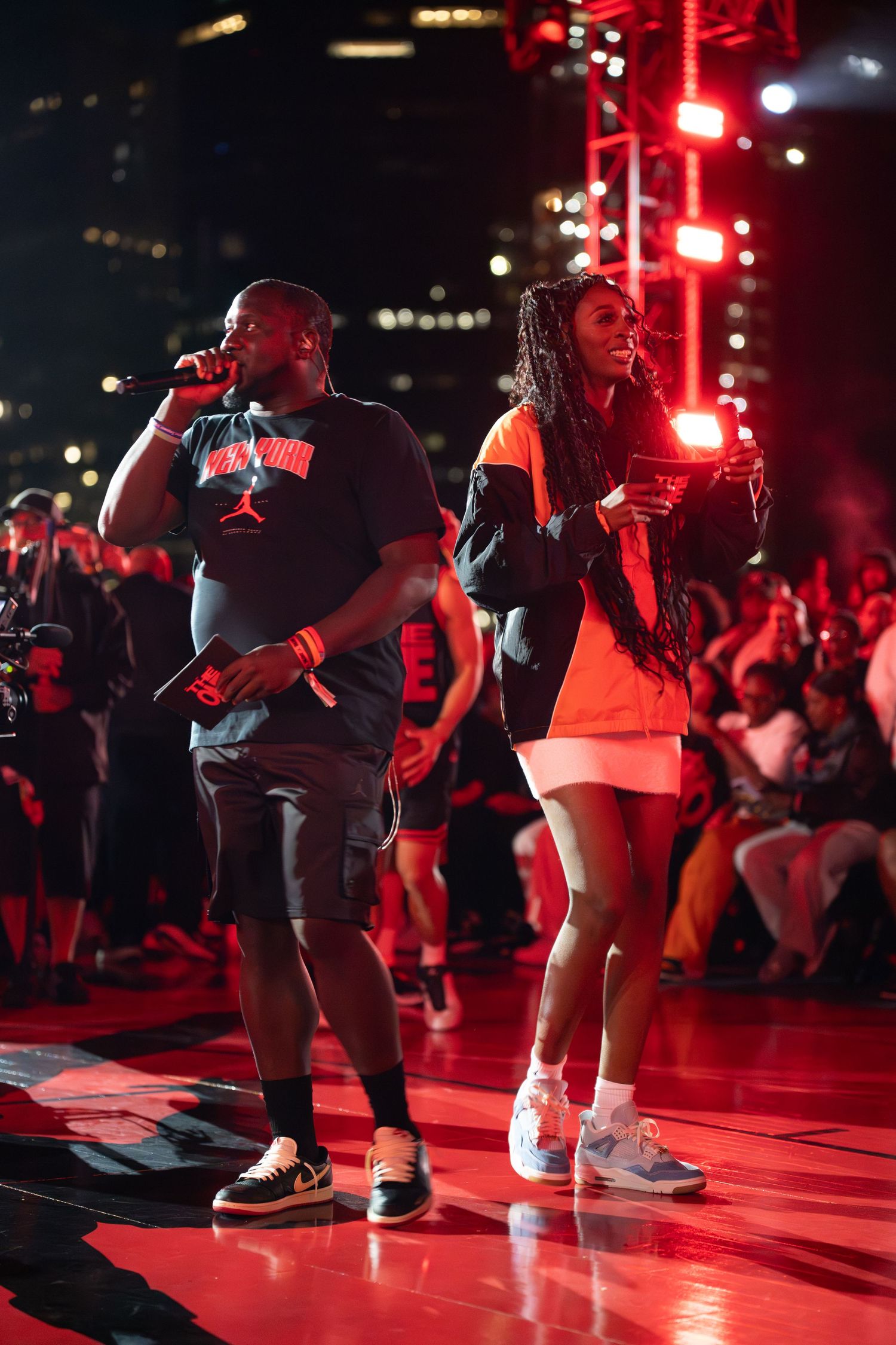 Two performers on stage at a nighttime concert with red lighting and an energetic crowd in the background.