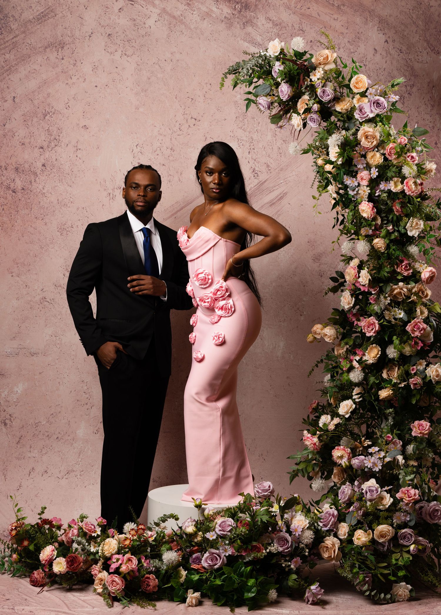 Romantic couple photoshoot with pink gown and black suit among floral arrangements.