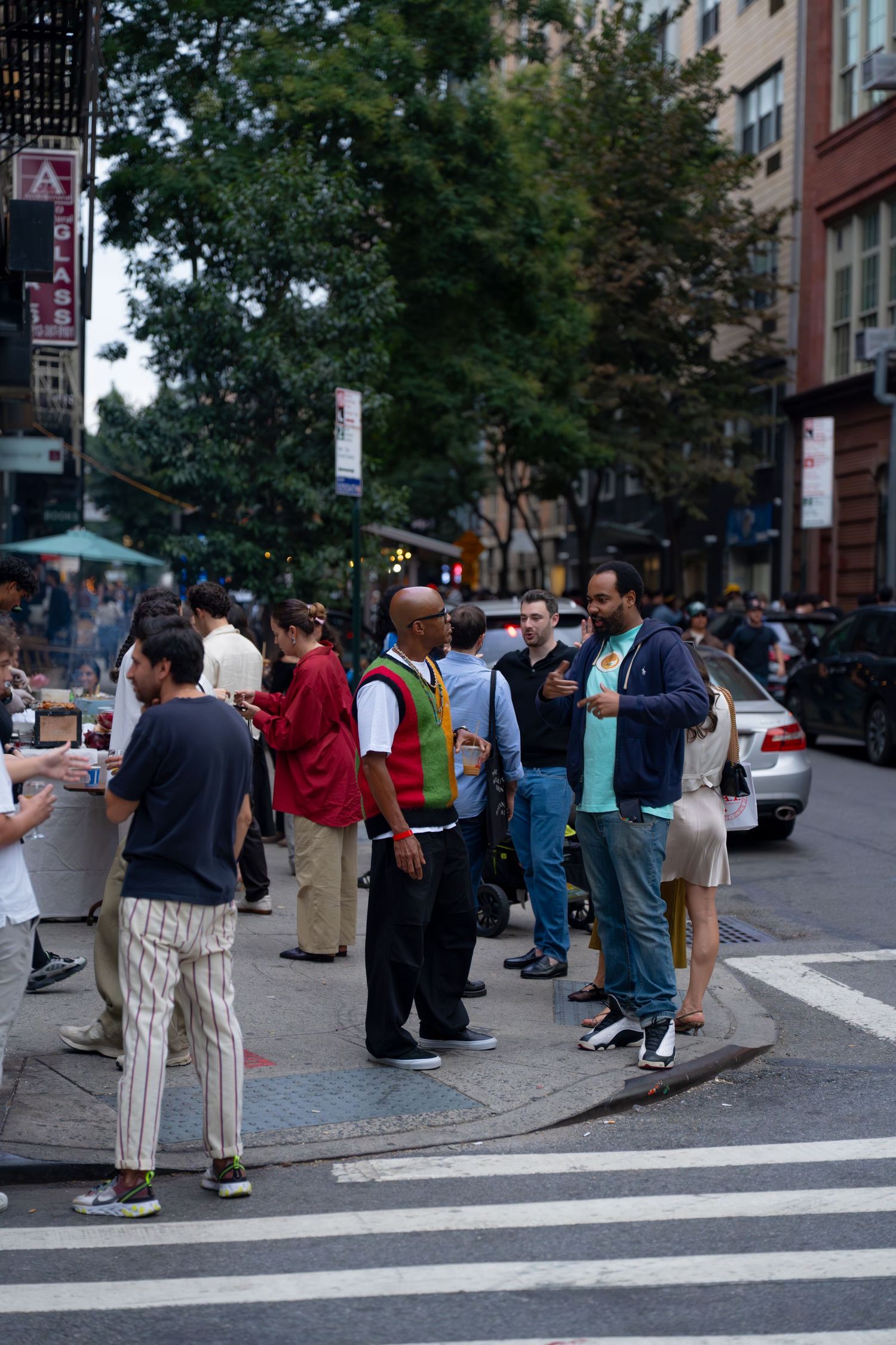 Groups of people gathered on an urban street corner with apartment buildings and trees visible.