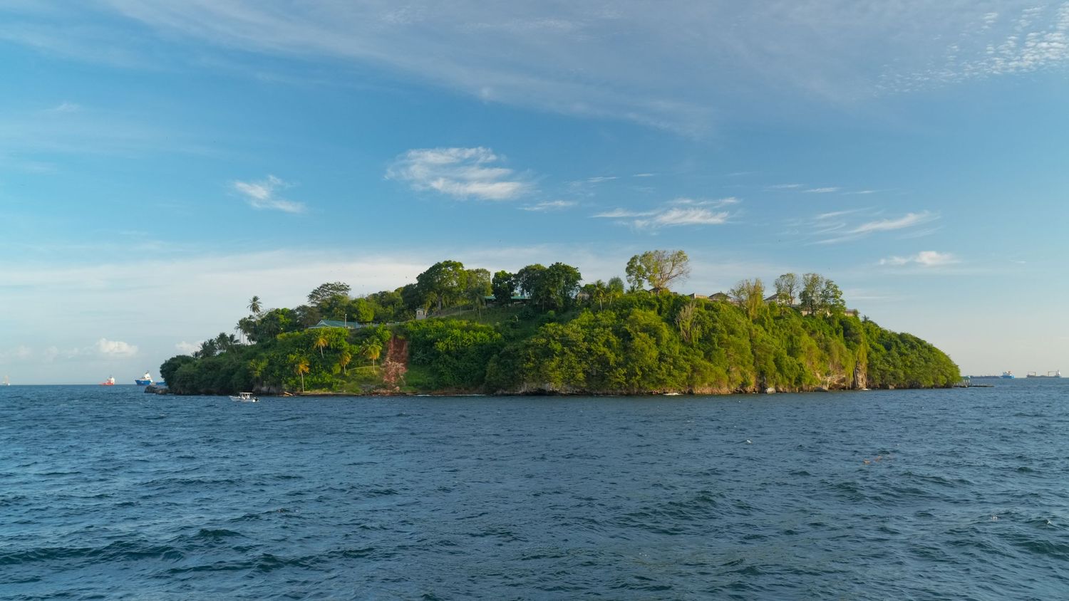 Small tropical island with lush green vegetation rises from blue ocean waters under bright sunny sky.