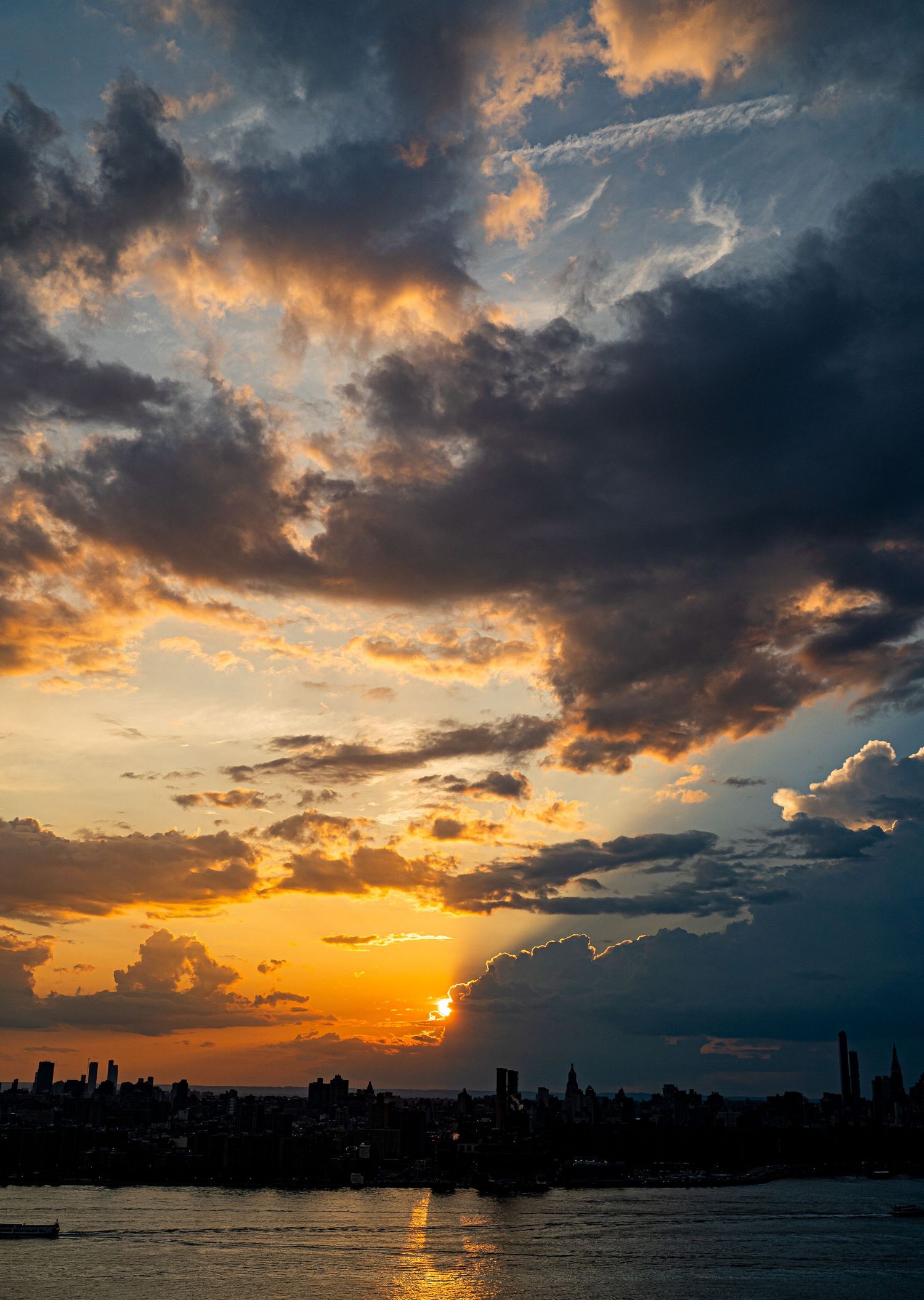 Dramatic orange sunset over city skyline silhouette with dark clouds and golden light reflecting on water.