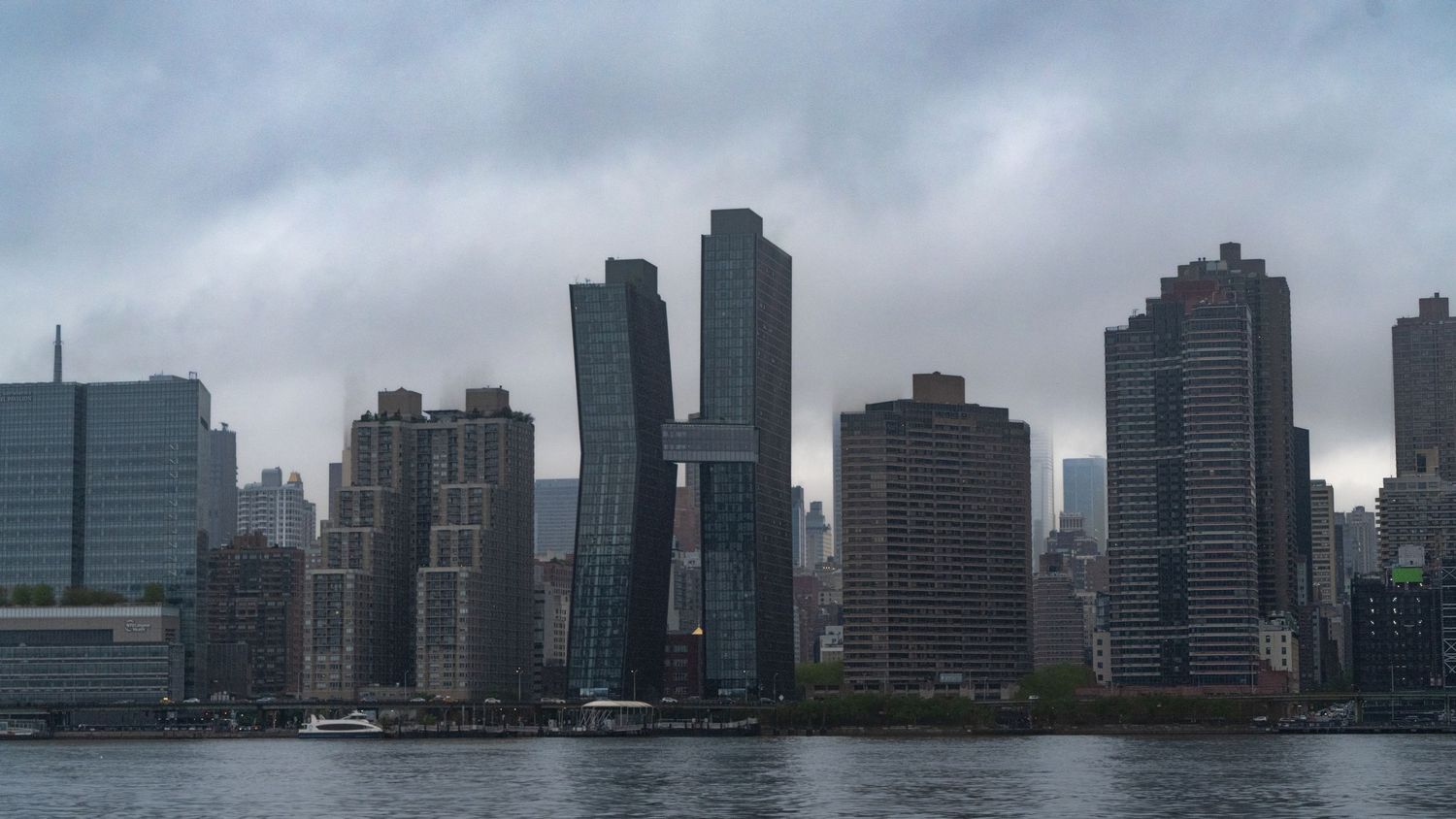Panoramic view of Manhattan skyscrapers under overcast skies reflecting in the East River.