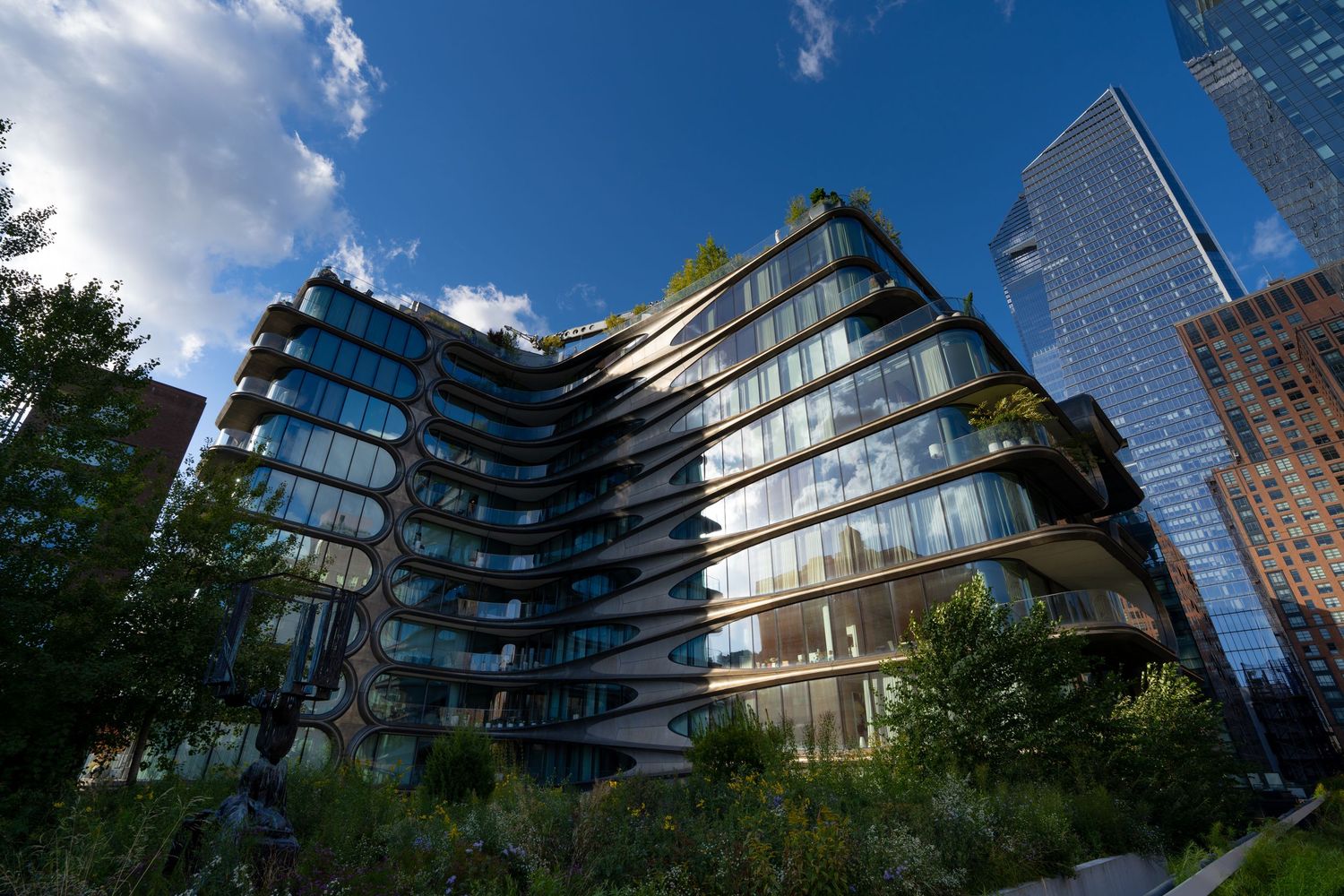 Modern curved glass luxury apartment building with green terraces against blue sky in urban NYC setting.
