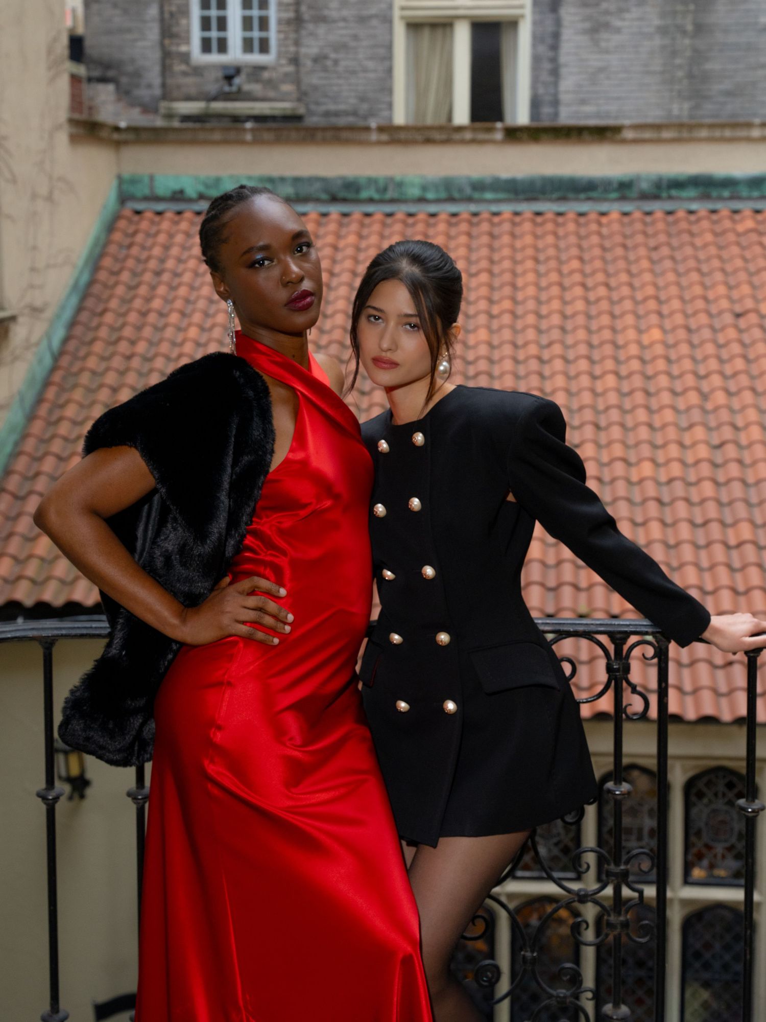 Two women pose on a balcony, one in a red satin gown with fur wrap, another in a black blazer dress with gold buttons.