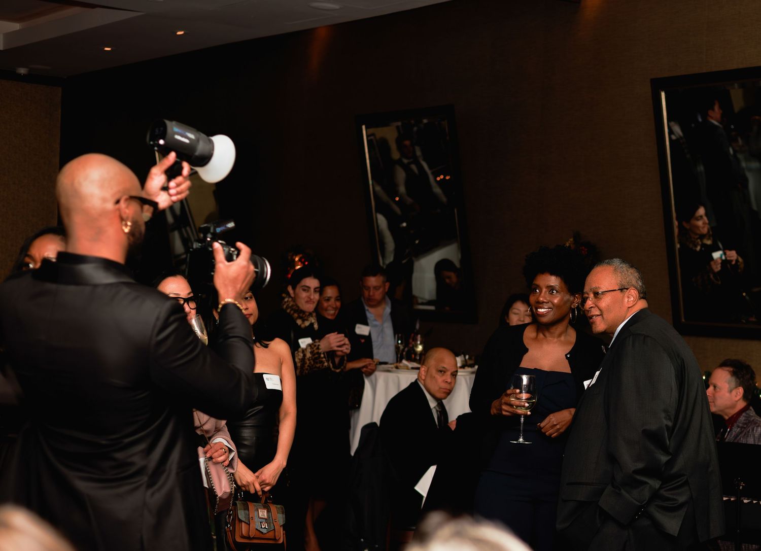 Photographer captures candid moment at formal evening event with guests mingling and holding drinks in dark room.