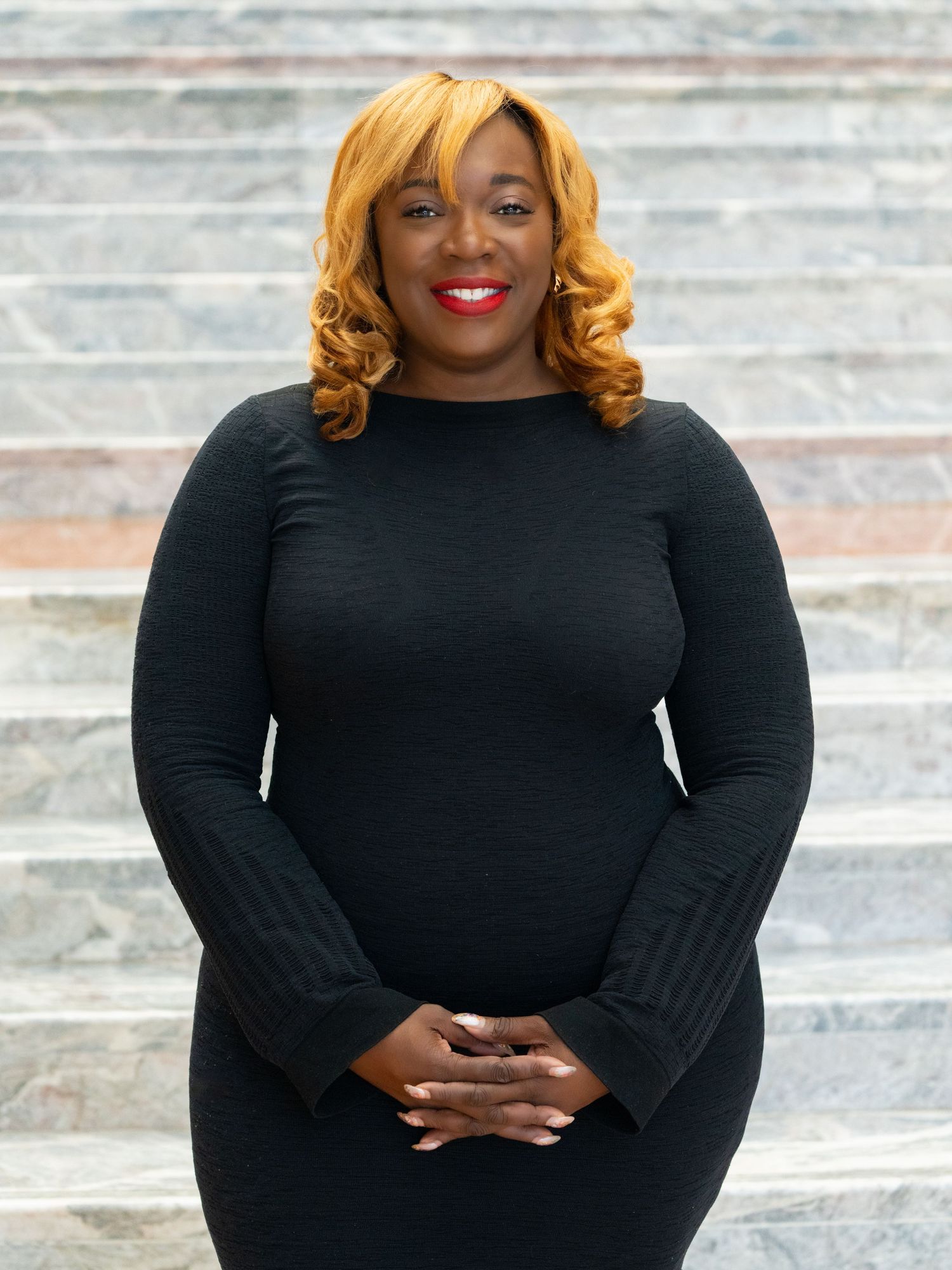 Professional headshot of a person in a black top with blonde curled hair against a light background.