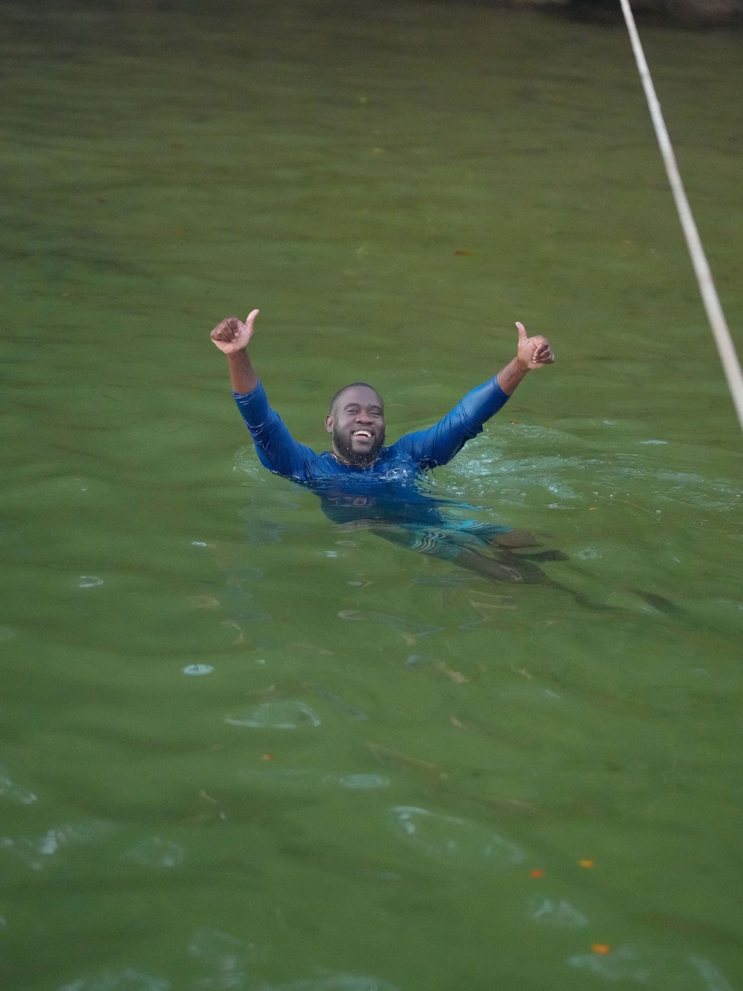 A person in a blue wetsuit raises their arms triumphantly while floating in murky green water.