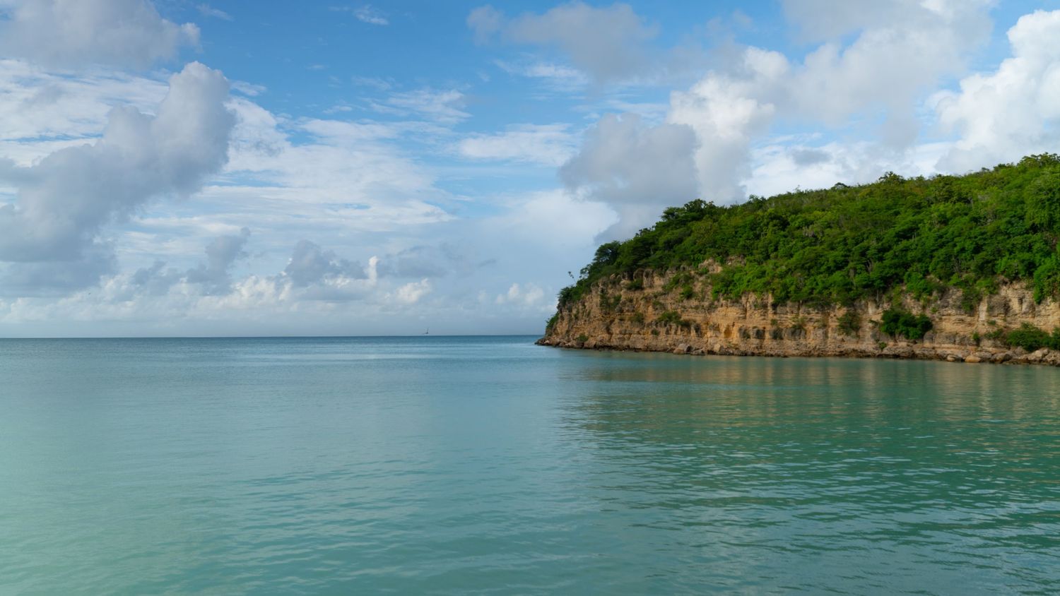 Panoramic view of a tropical coastline with turquoise waters, green cliffs and puffy white clouds in the blue sky.
