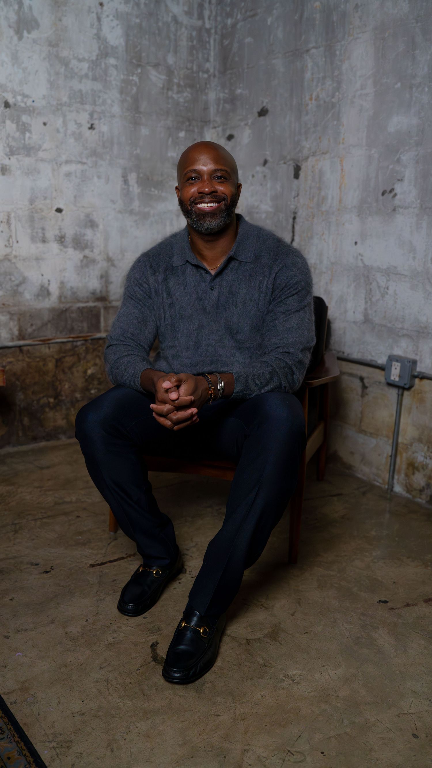 Person in dark denim outfit sits smiling on concrete floor in industrial space.