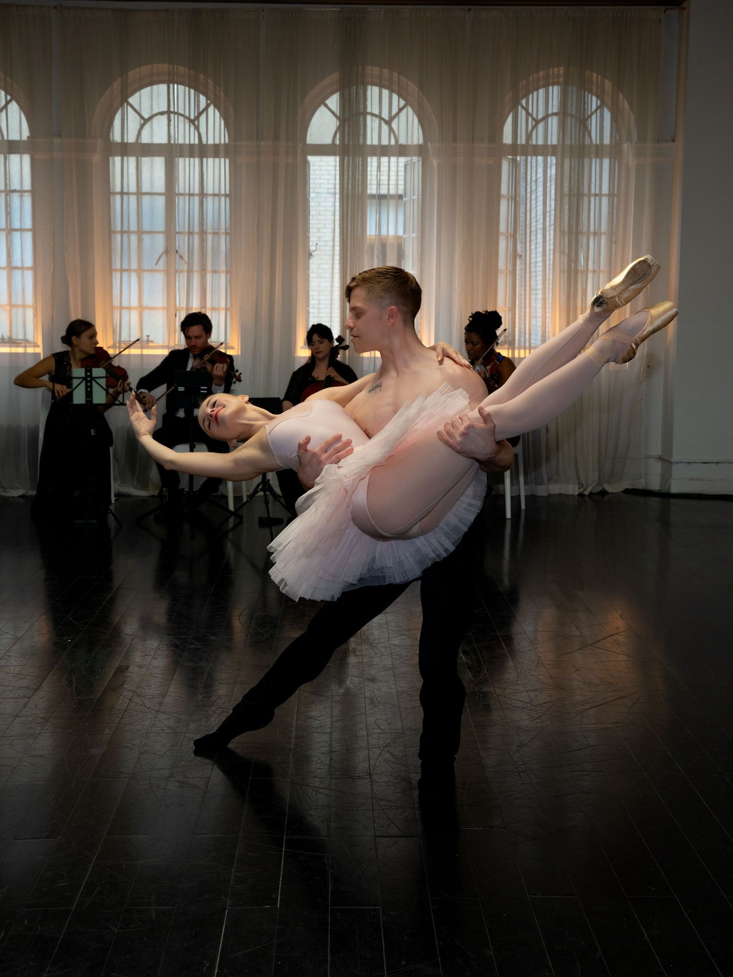 Male ballet dancer lifts female ballerina in white tutu during elegant performance with live musicians in background.