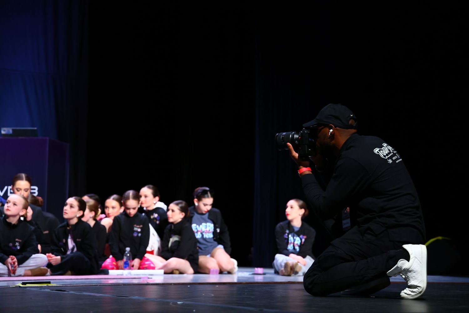 Photographer kneeling on stage capturing young dancers in black outfits seated in a row during a dance performance event.