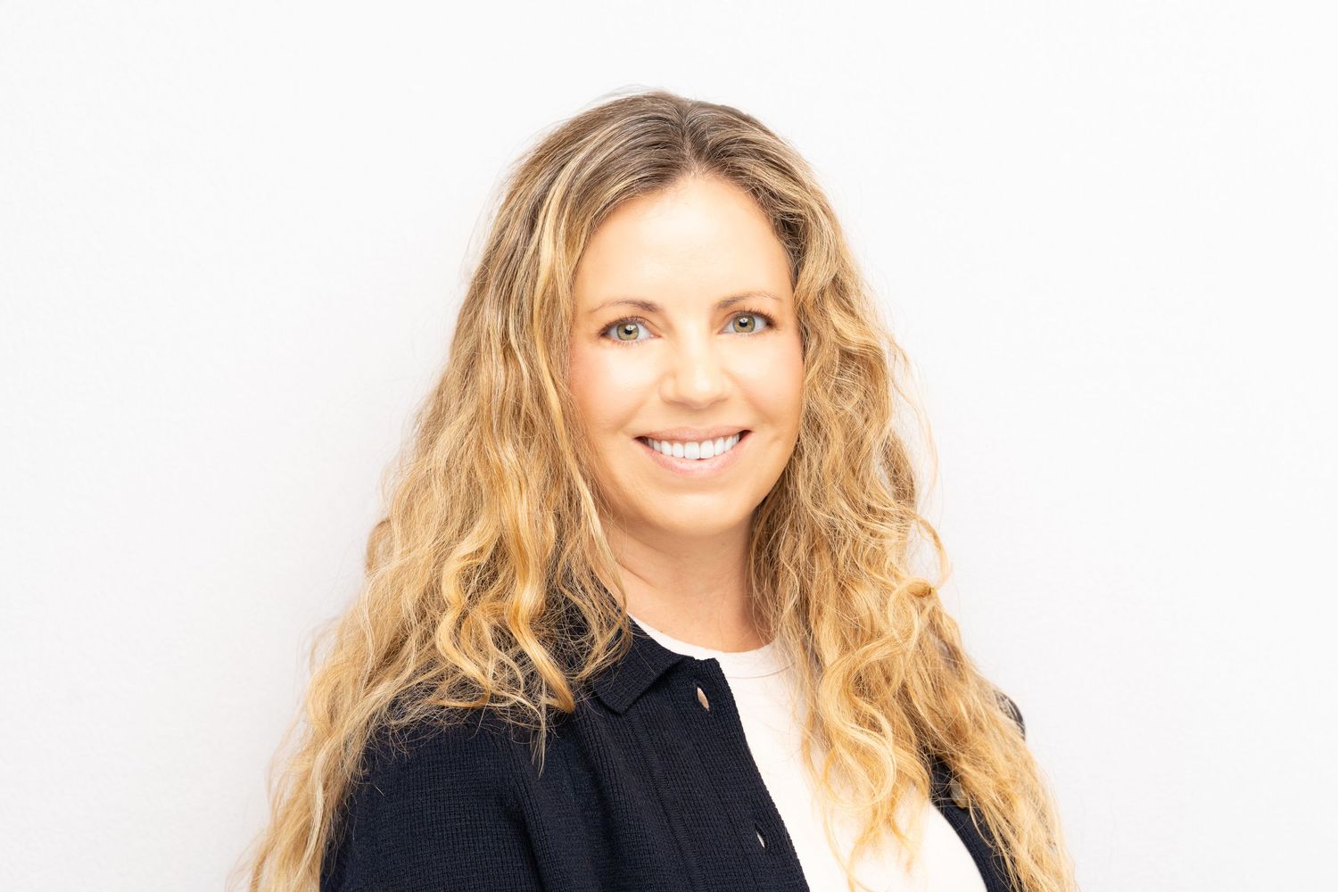 Professional portrait of a person with long blonde wavy hair wearing a black and white outfit against white background.