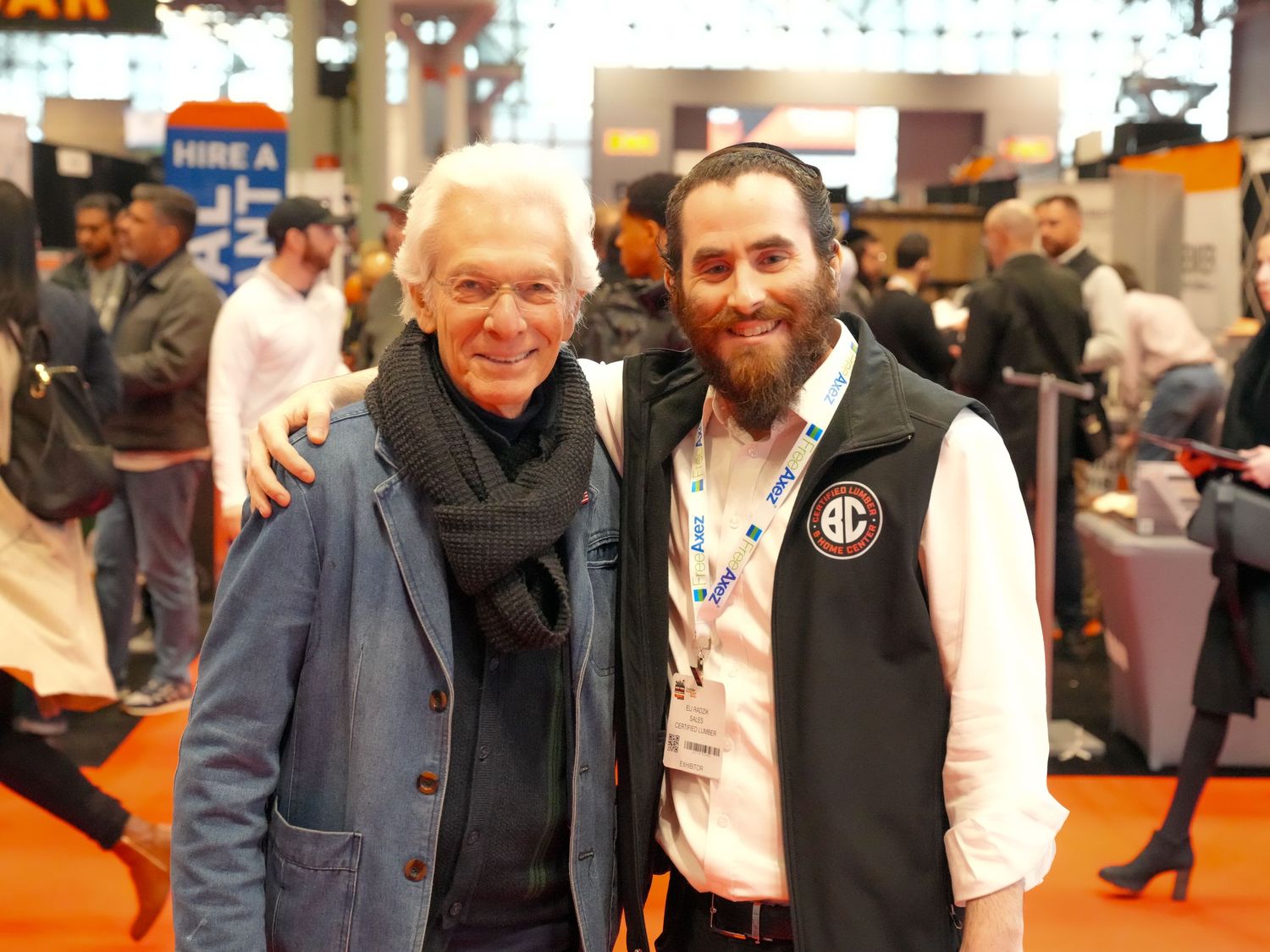 Two men smiling and posing together at a busy indoor trade show or convention event.