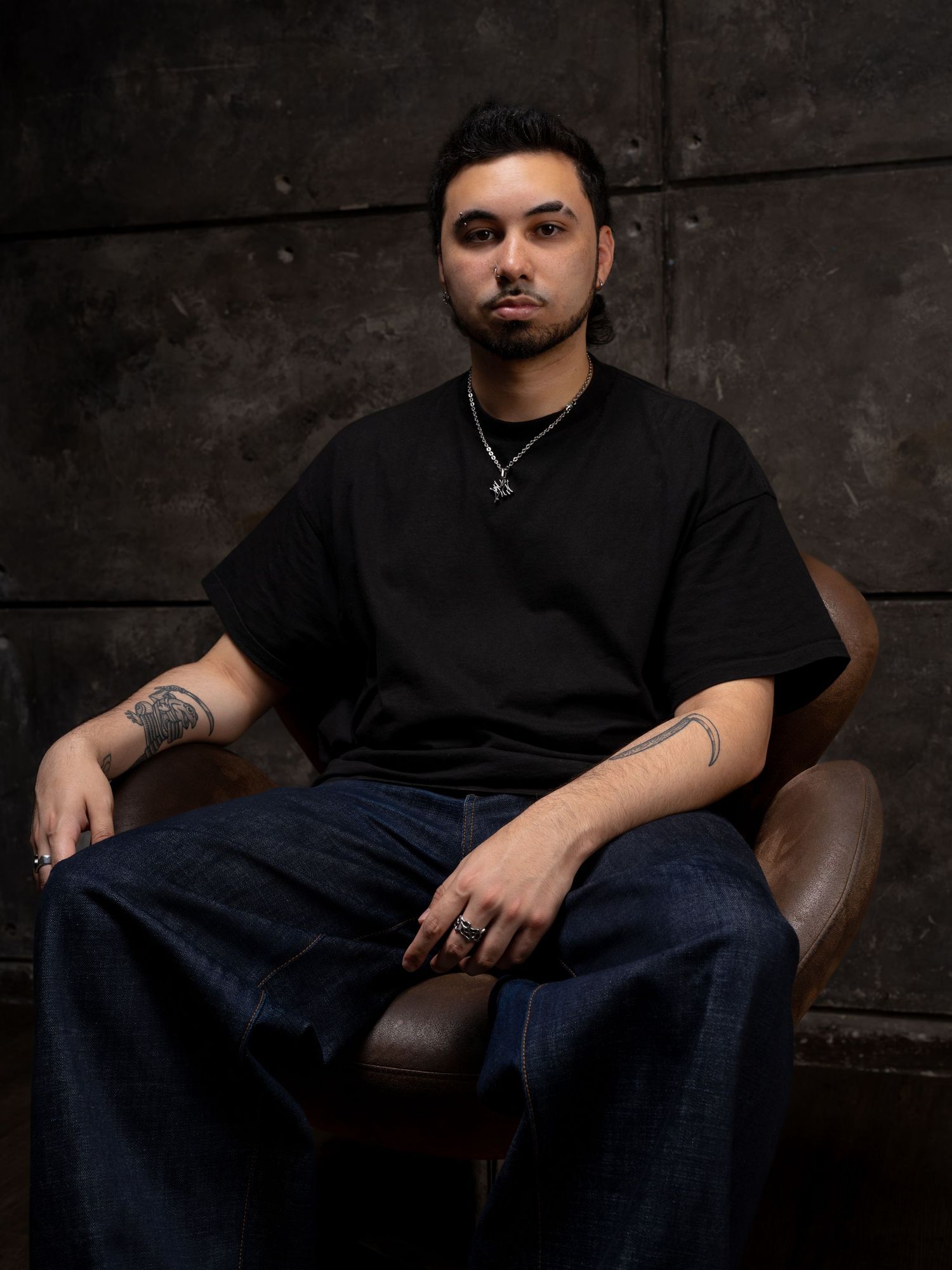 Young man with tattoos sits in leather chair wearing black tee and jeans, posing against dark textured wall.