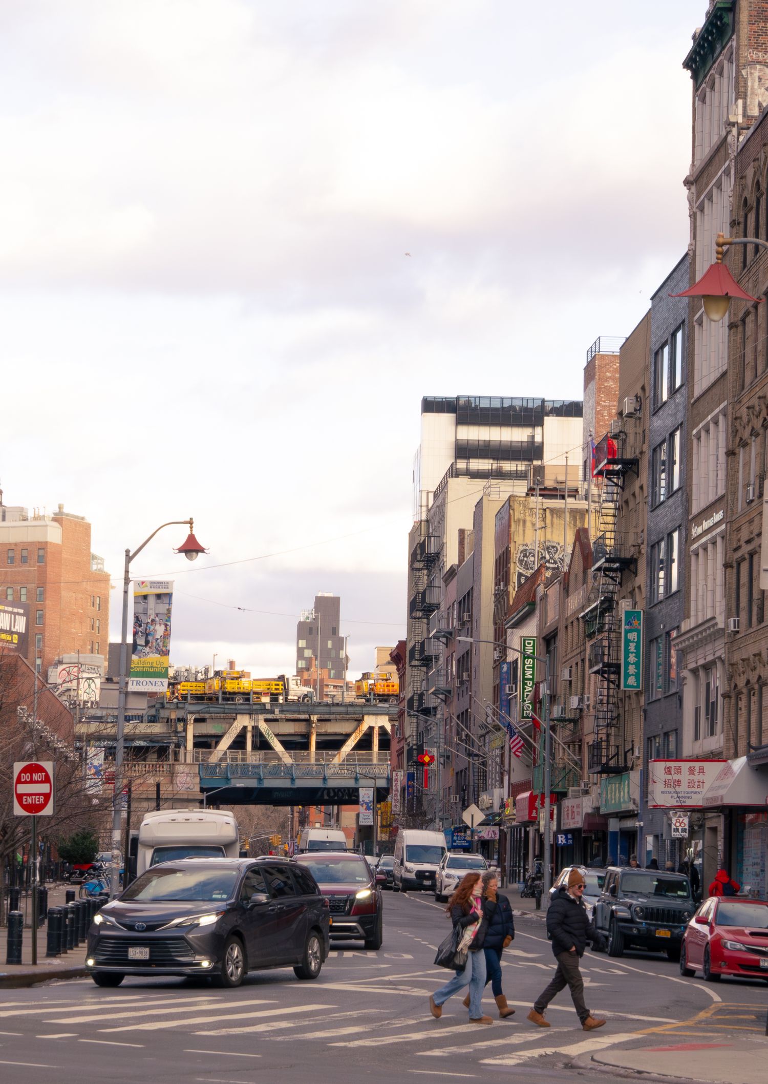 Urban street scene in Brooklyn with brick buildings, parked cars, and pedestrians crossing under cloudy skies.