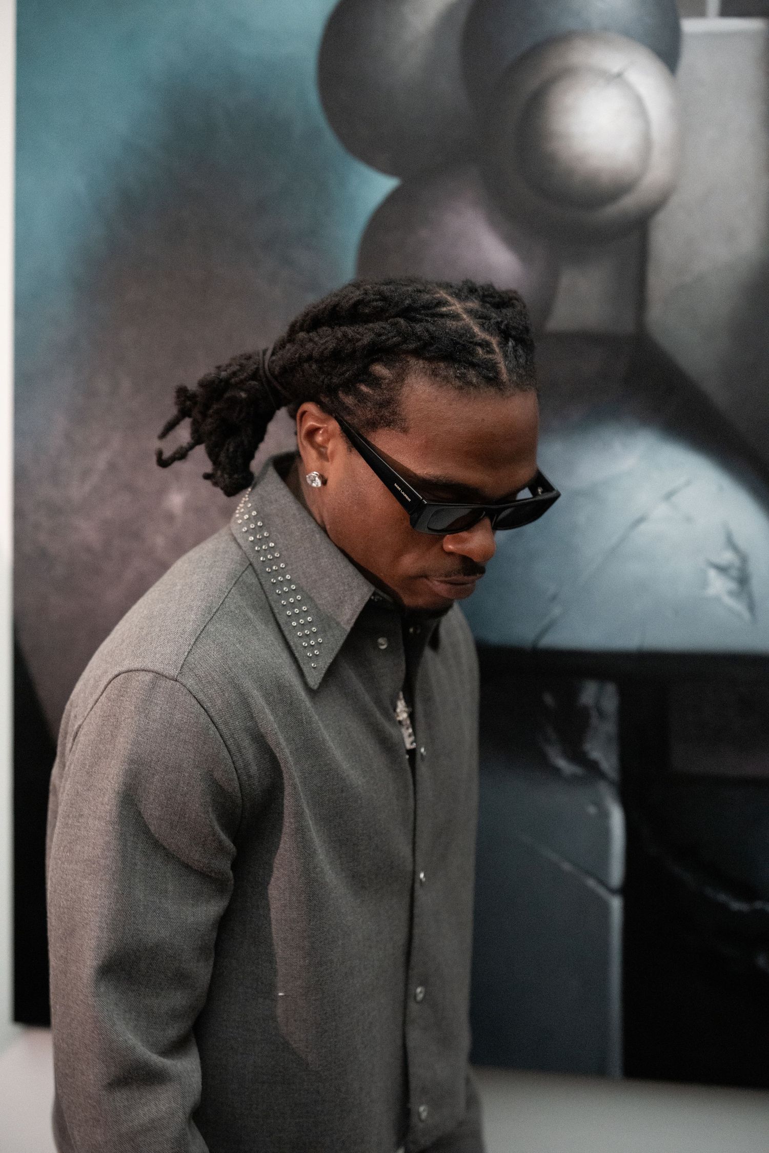 Figure in grey collared jacket and sunglasses shown in profile against abstract background.