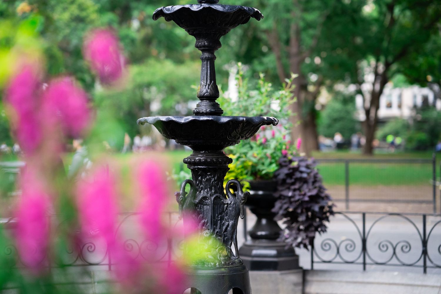 Ornate black metal fountain in a garden with pink flowers blurred in the foreground.