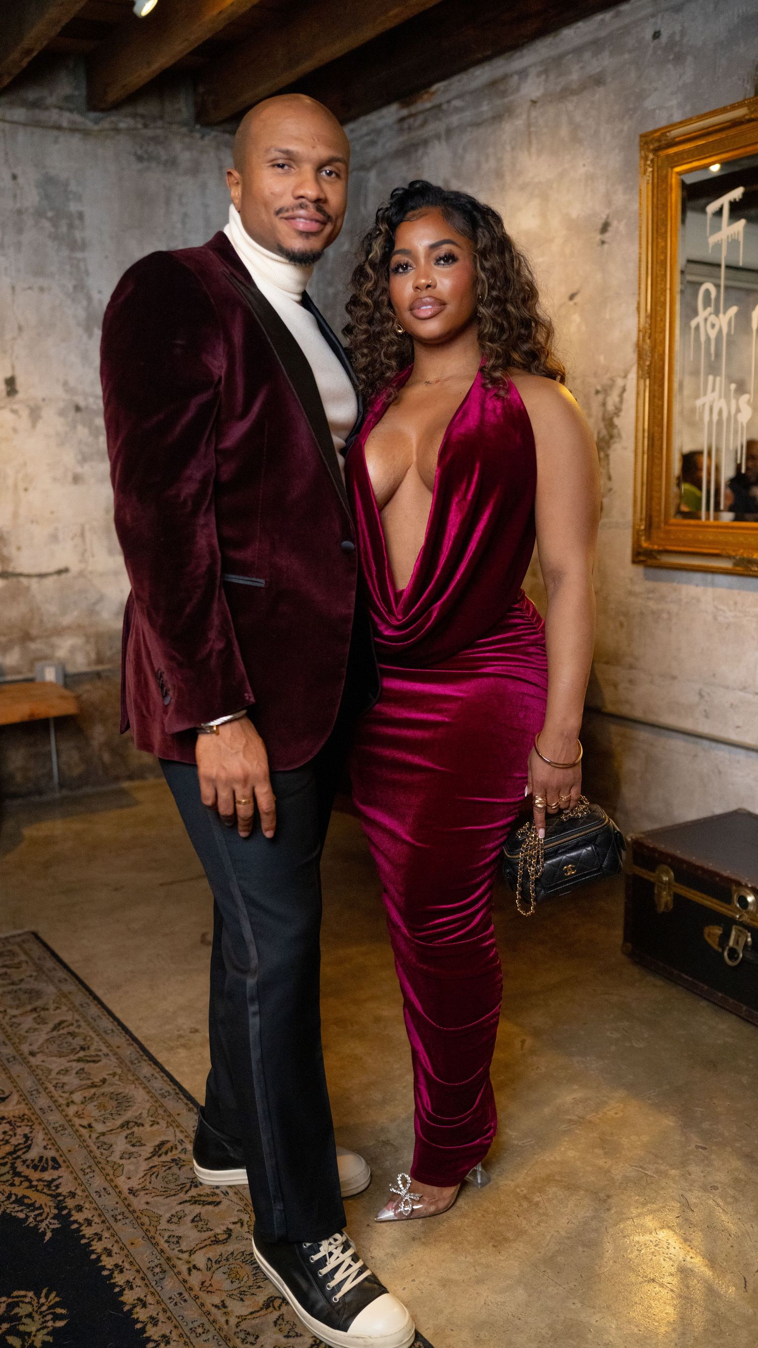 A couple in elegant burgundy velvet attire poses together in an industrial-style space with exposed walls and wooden beams.
