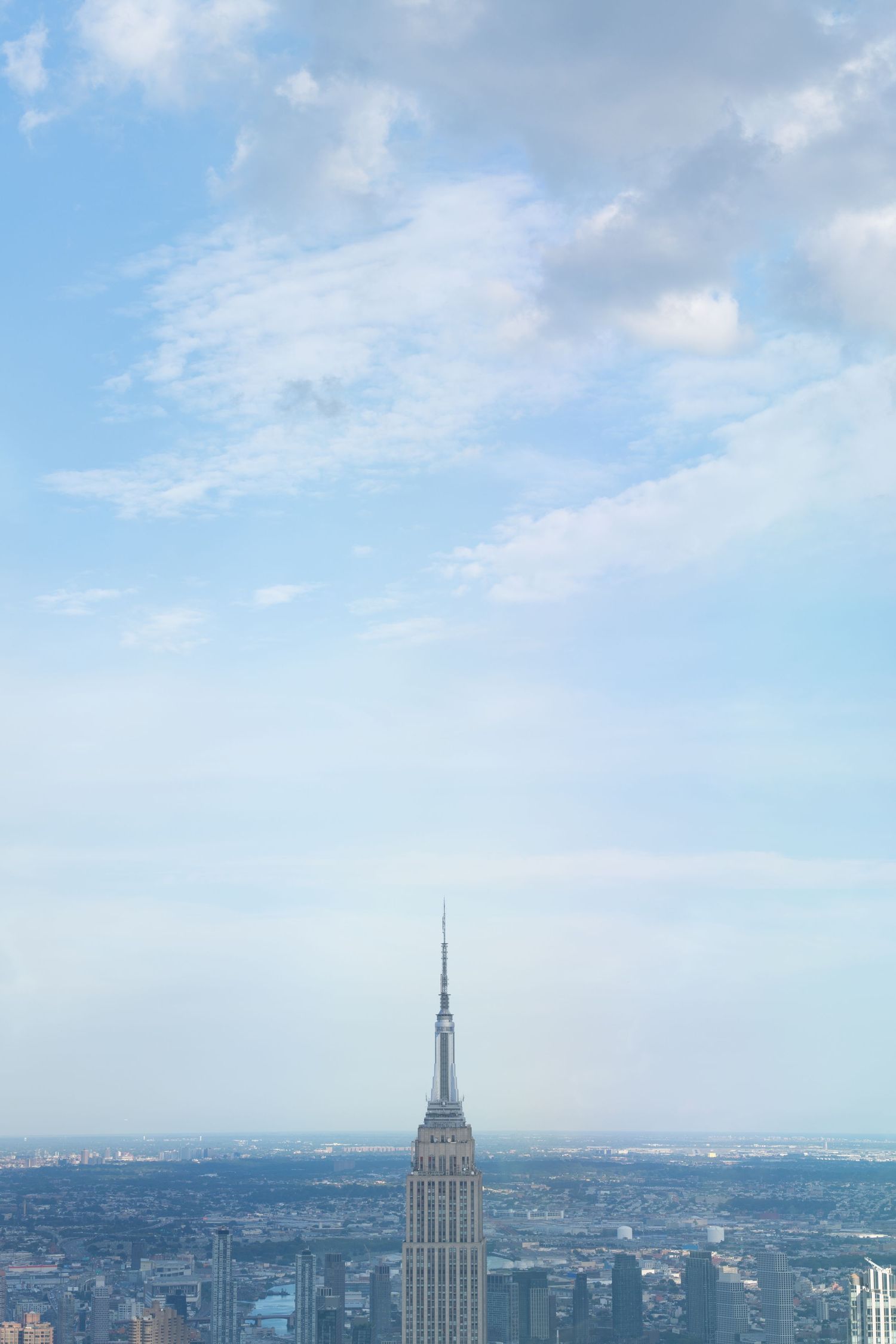 Empire State Building rises above Manhattan skyline against bright blue sky with white clouds.