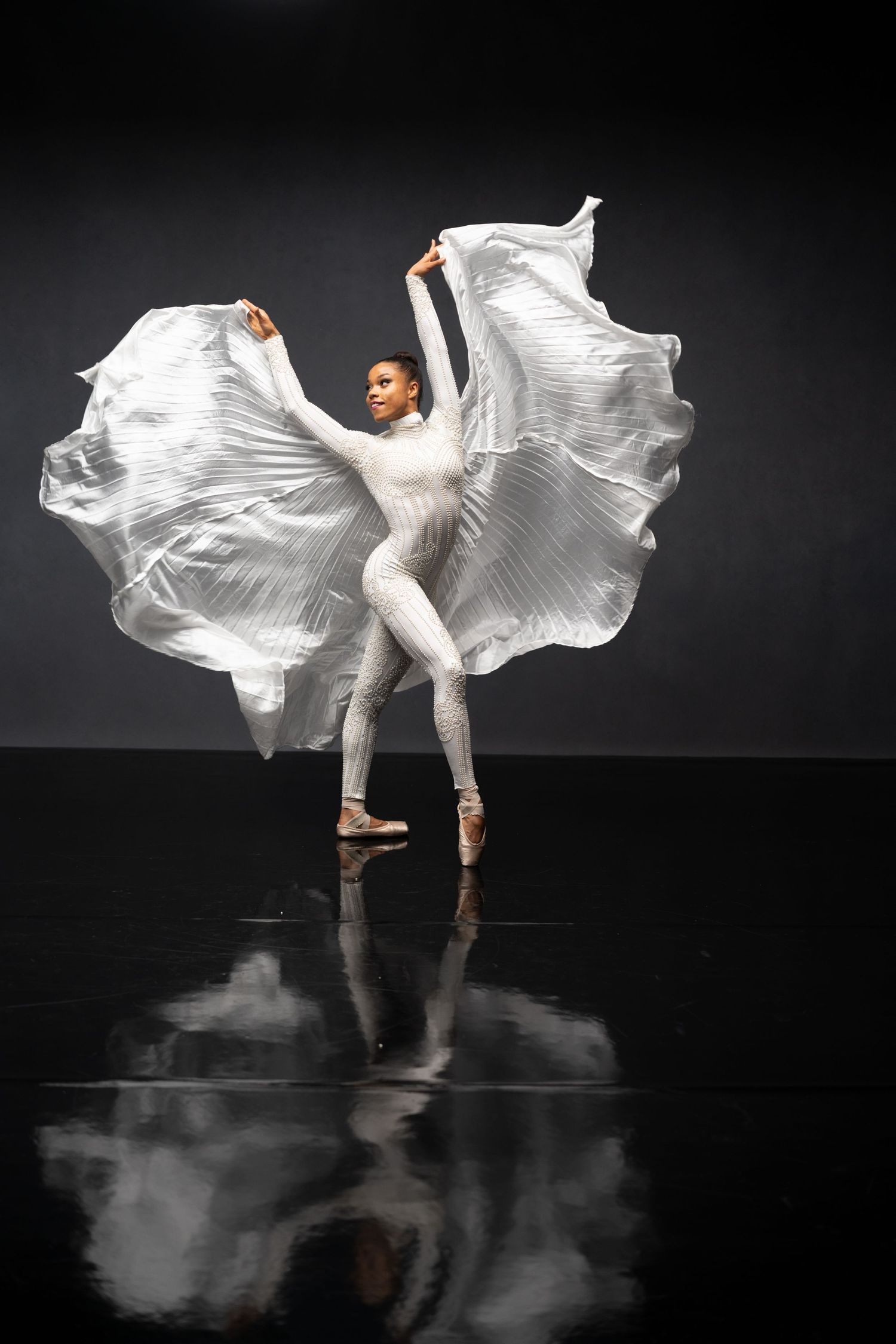 Dancer performs with billowing white fabric creating ethereal shapes on dark reflective stage.