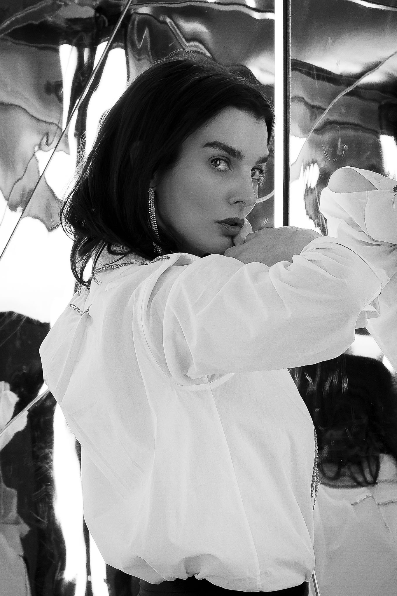 Black and white portrait of a young woman in a white oversized jacket posing with arms raised near reflective surfaces.