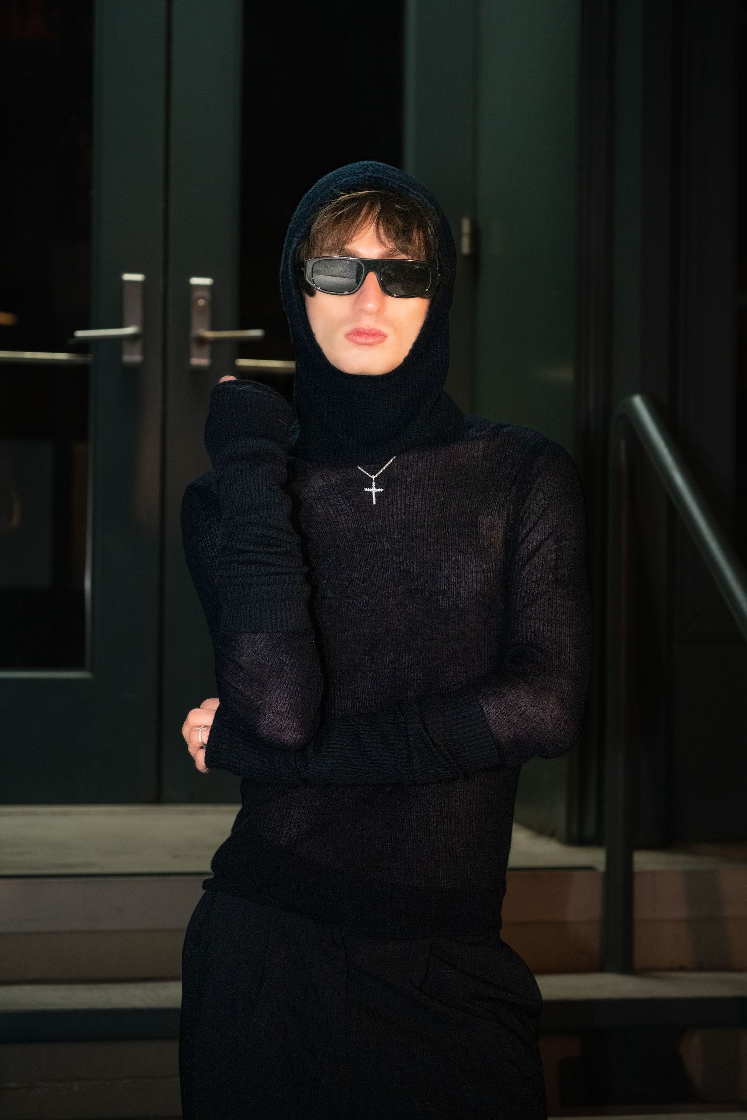 Person in dark clothing and sunglasses sitting in dramatic low-light setting.