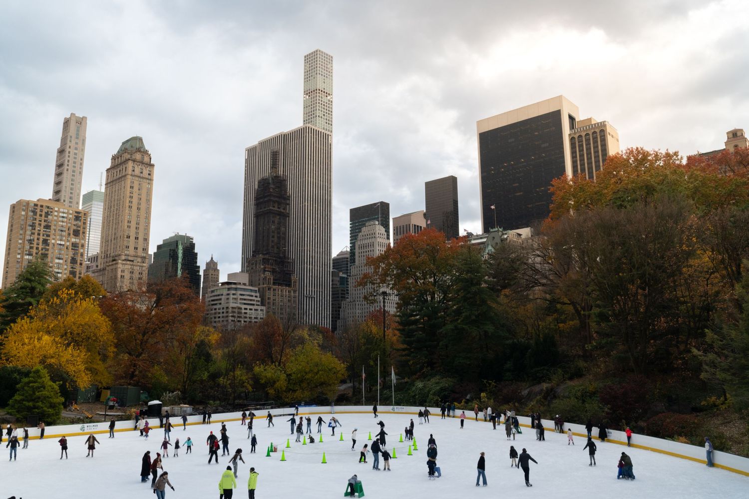 A panoramic view of people ice skating at a rink in Central Park with Manhattan skyscrapers in the background.
