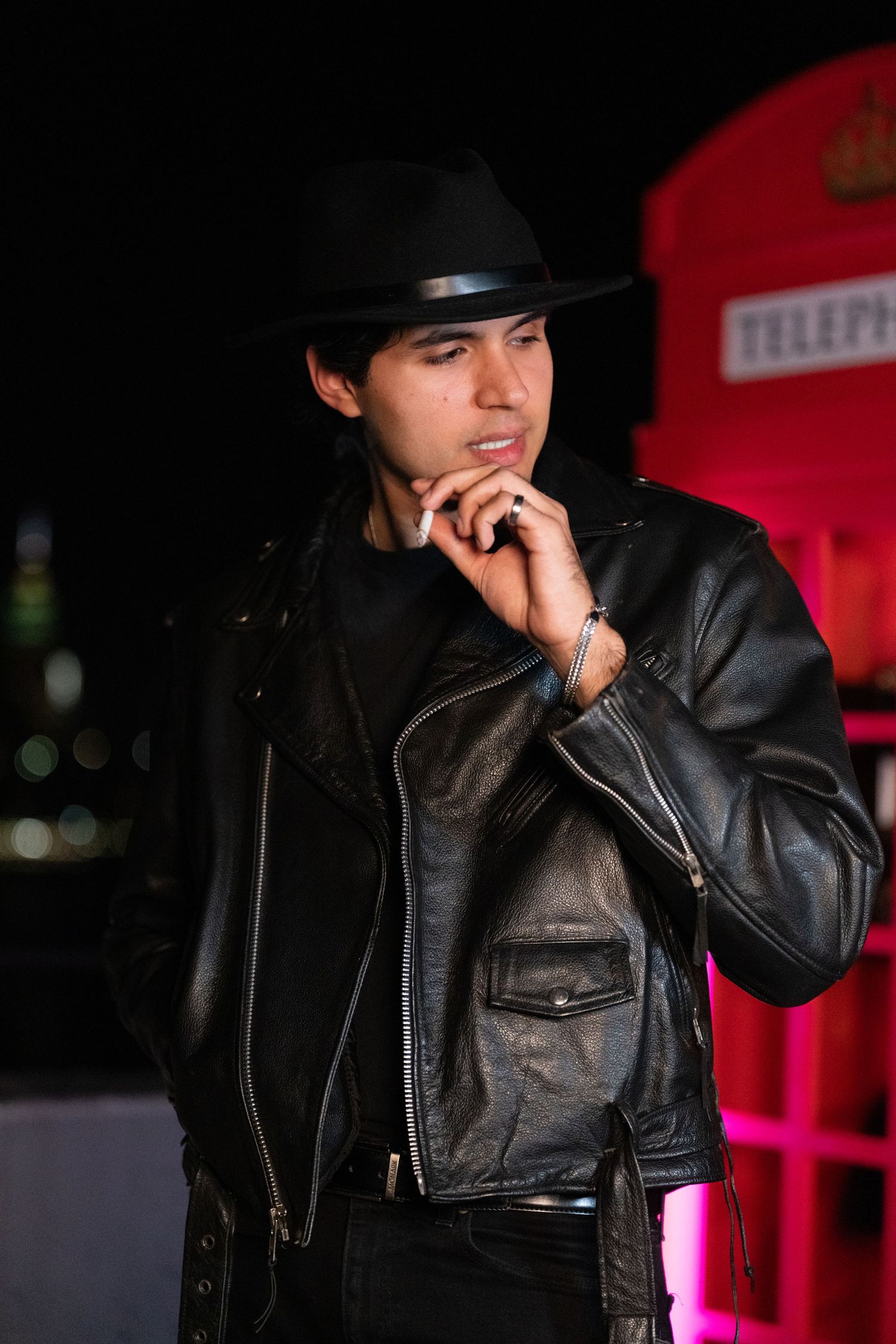 Figure in black leather jacket and hat poses thoughtfully in dark setting.