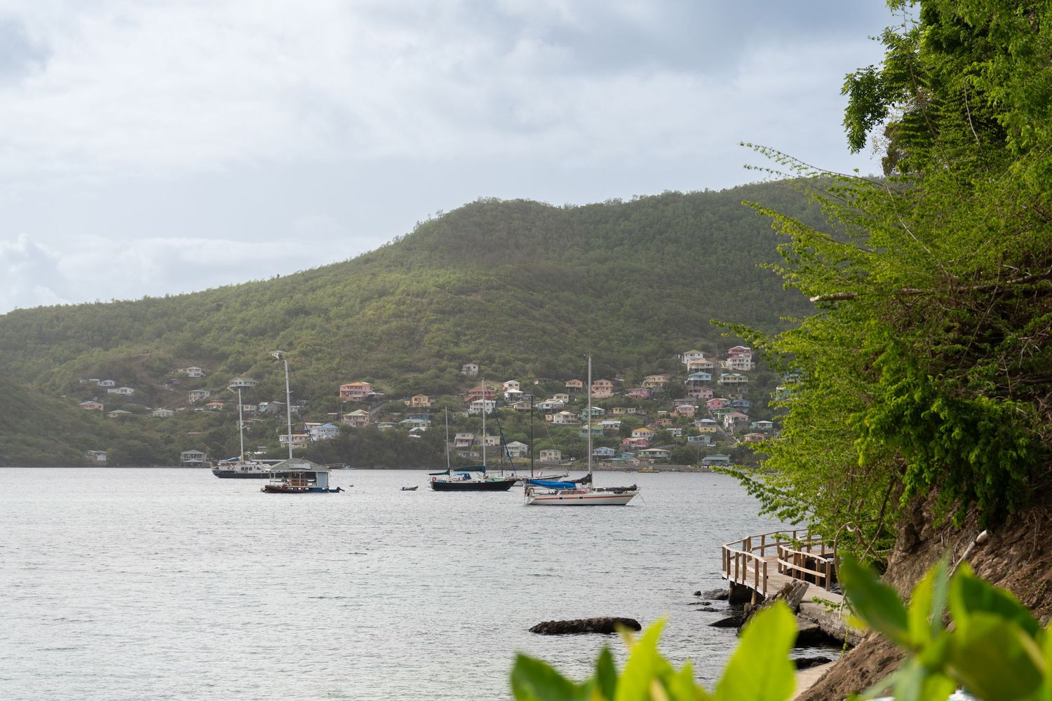 Coastal harbor view with boats anchored near mountainous shoreline under cloudy skies.
