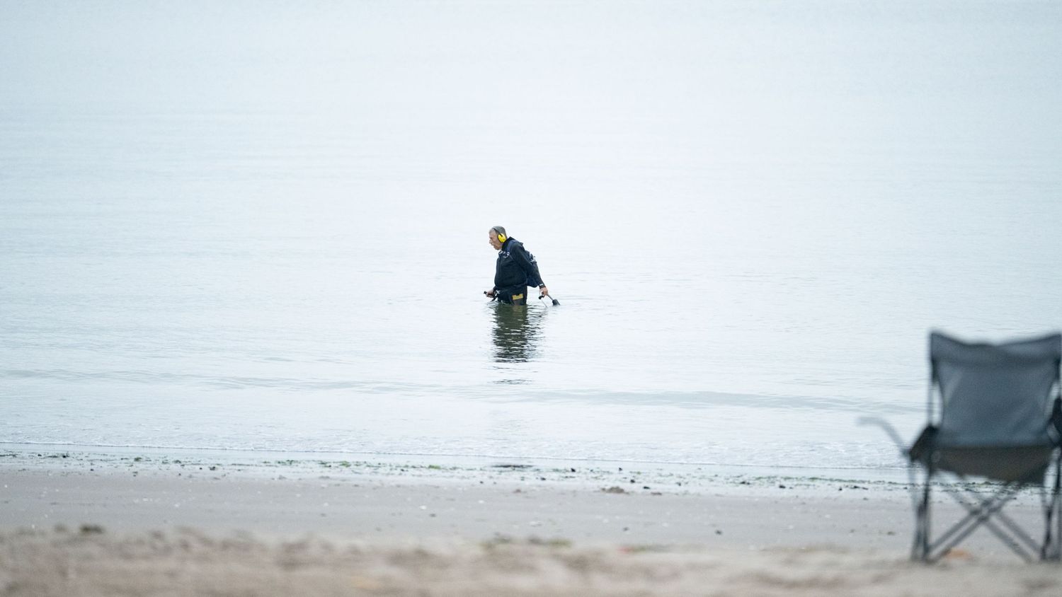 Lone figure wading in misty waters with an empty beach chair in the foreground.