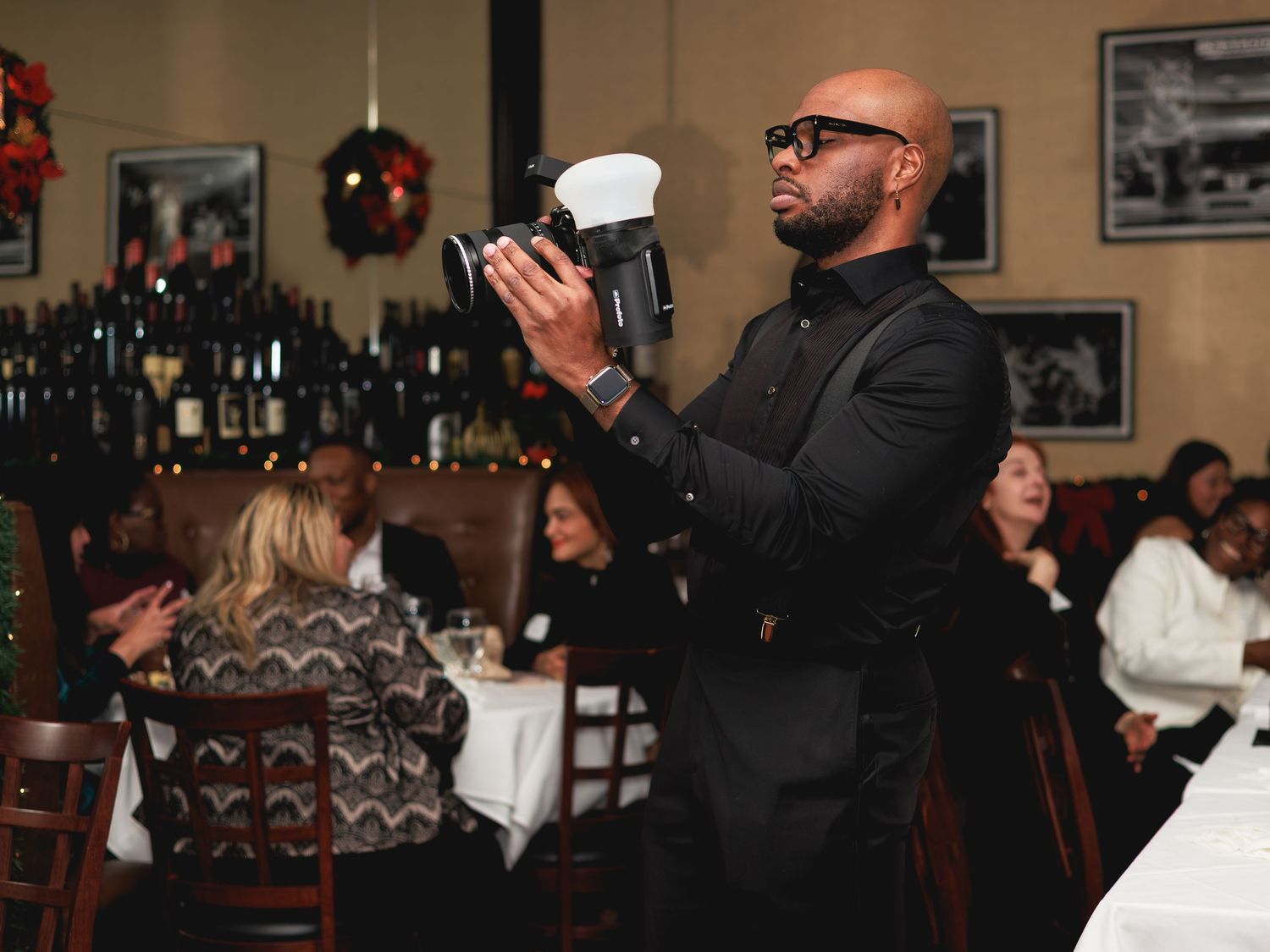 A person in a black turtleneck and glasses holds up a wine glass while standing in a restaurant bar area.