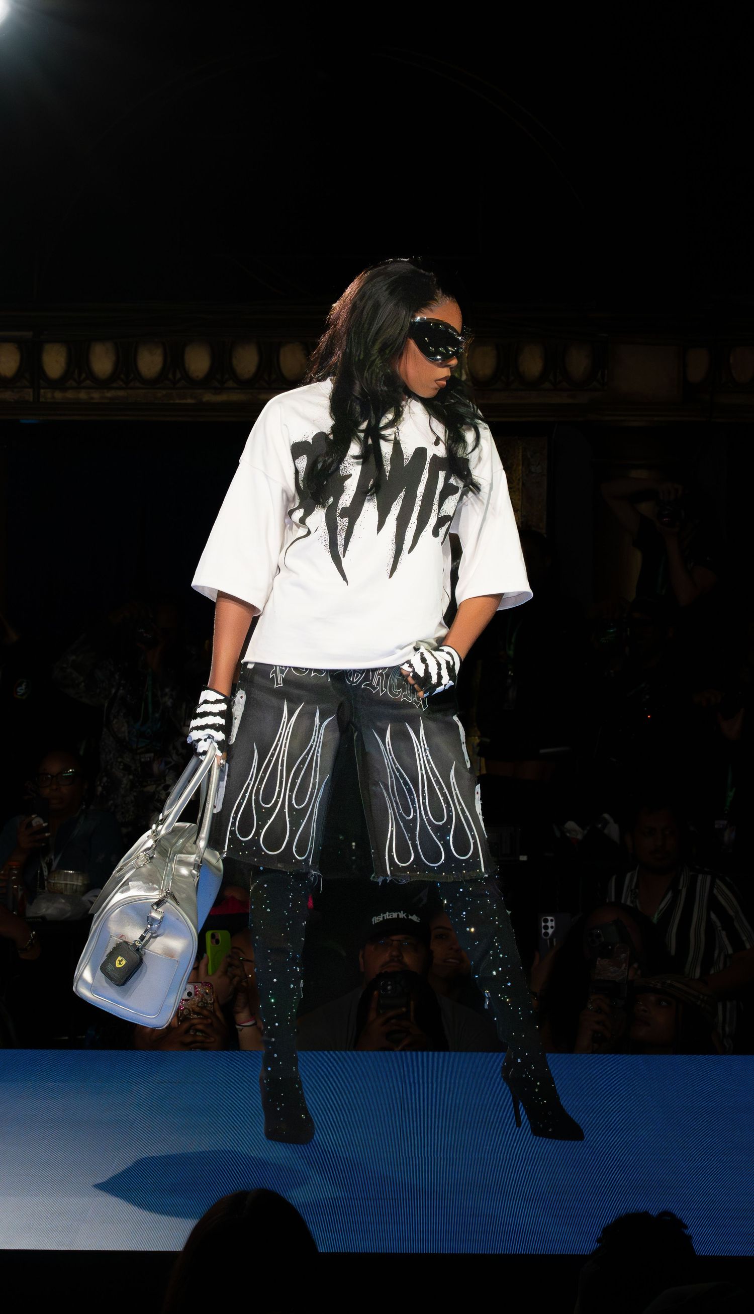 Model in white t-shirt and decorated black pants poses on blue-lit runway.