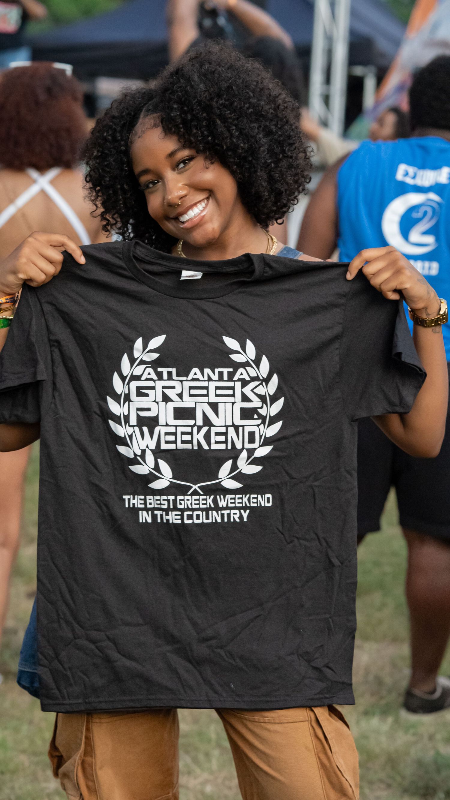 Person holding up black t-shirt with Greek Weekend logo and text at outdoor event.