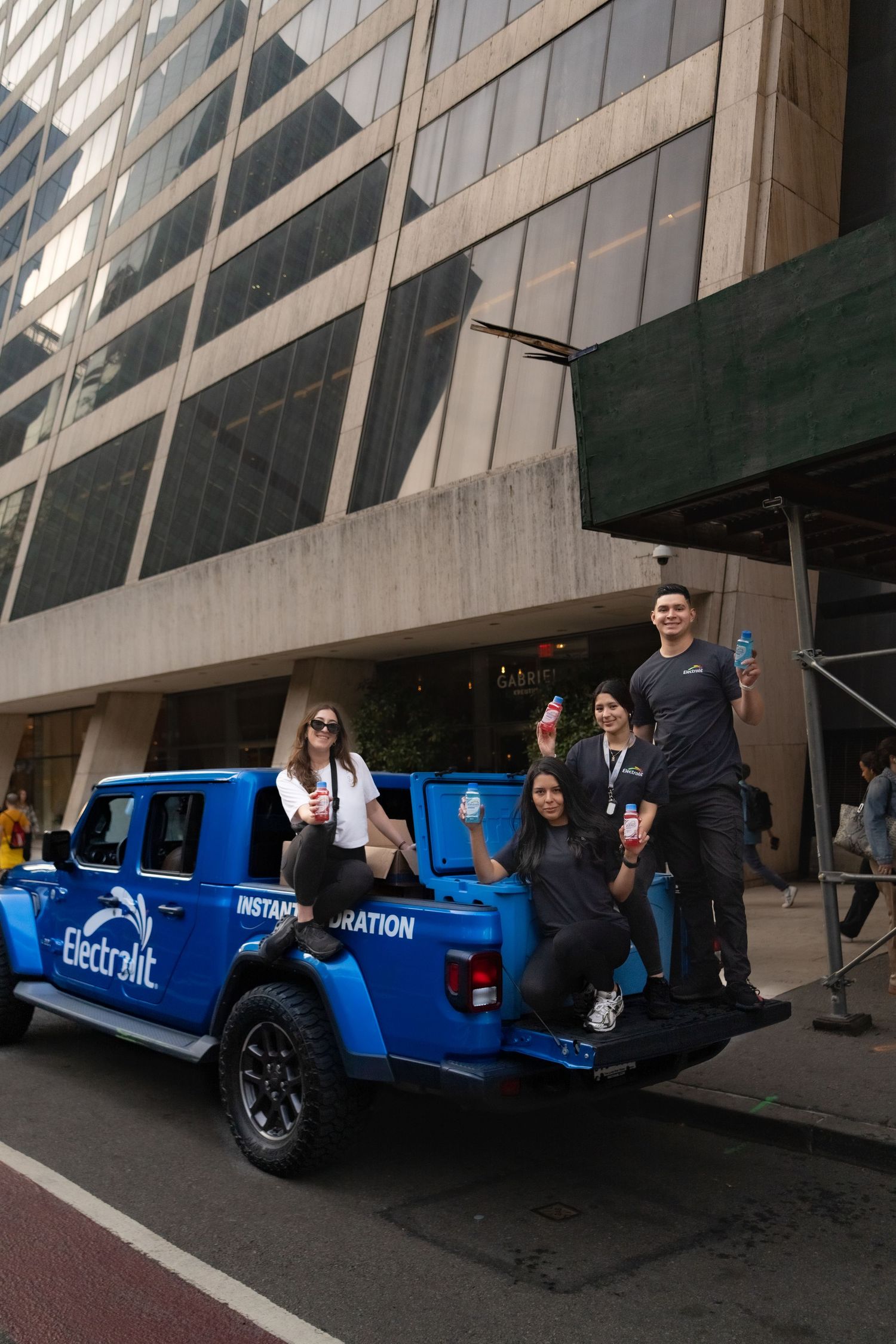People gathered around a bright blue promotional vehicle parked outside a modern glass building.