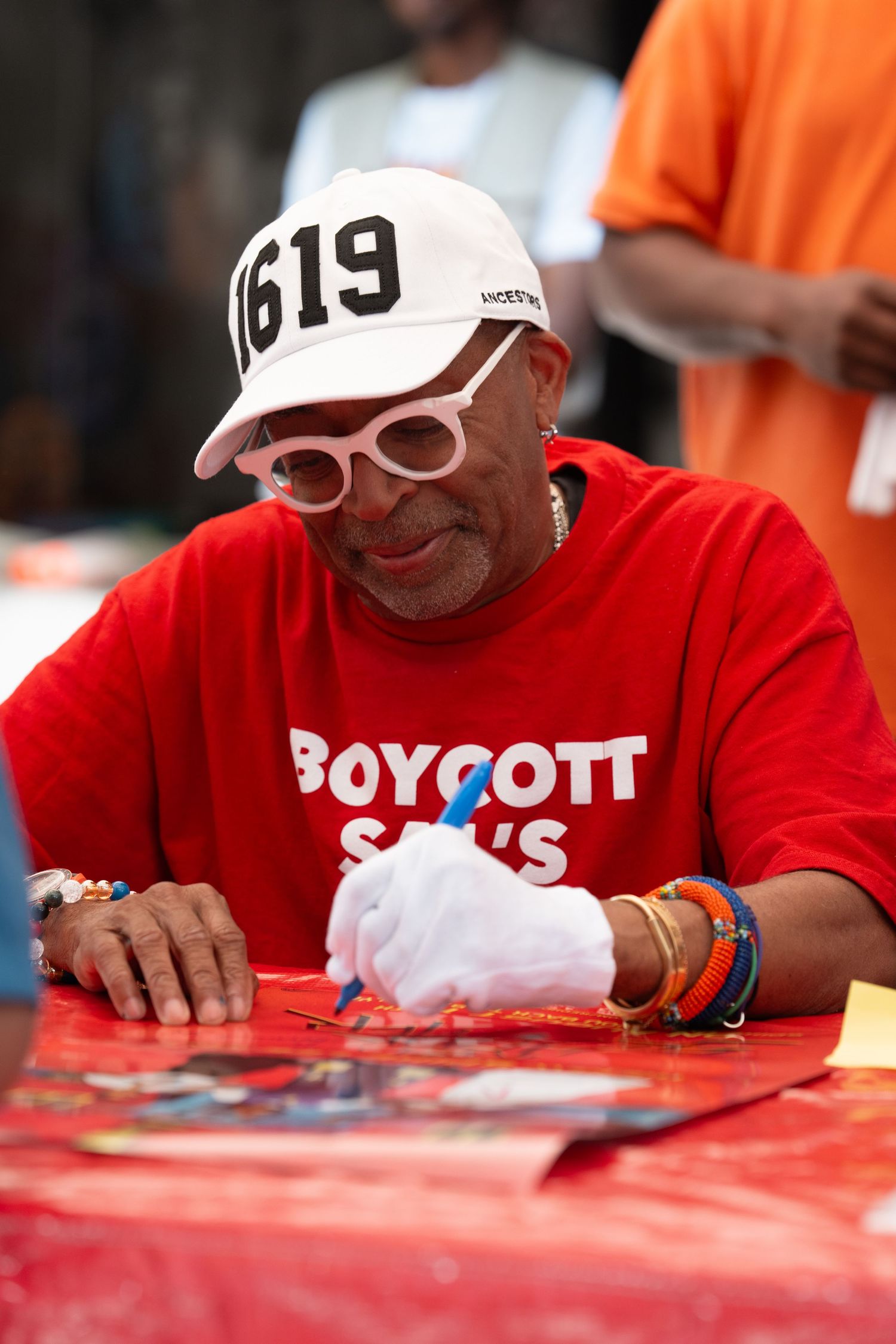 Person in red shirt and white cap signing autographs at outdoor event.