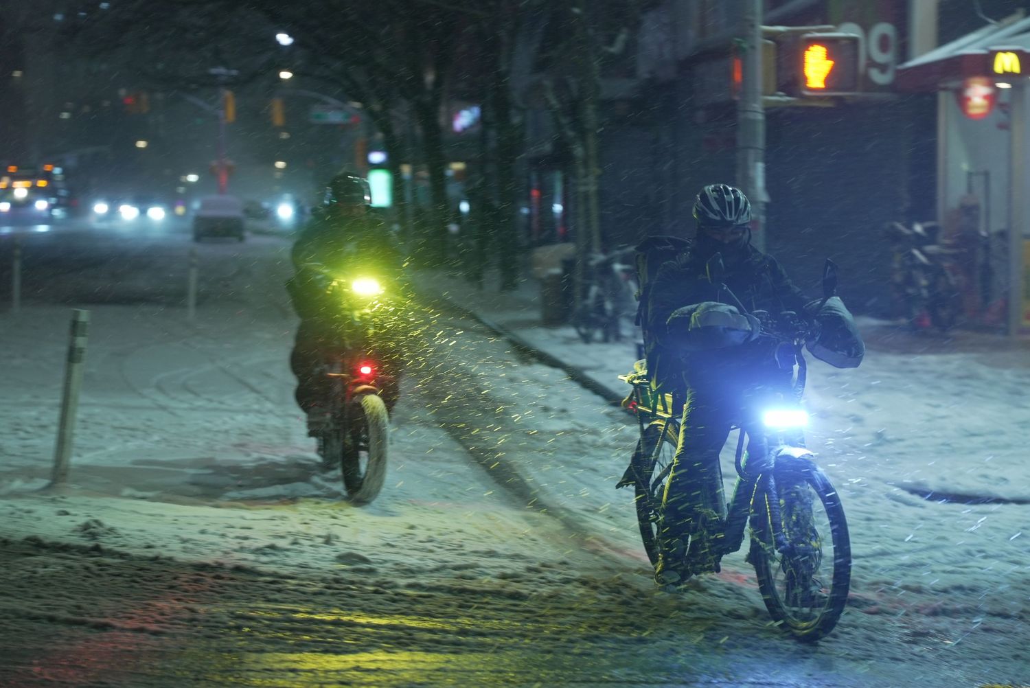 Cyclists ride through snowy city streets at night with bright lights illuminating their path in winter conditions.
