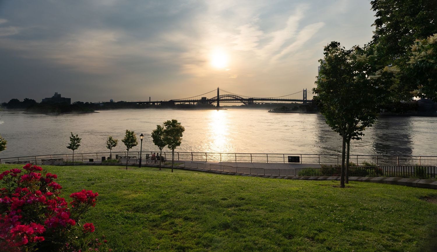 Sunset over a river with a bridge, green park, and red flowers in the foreground on a hazy evening.