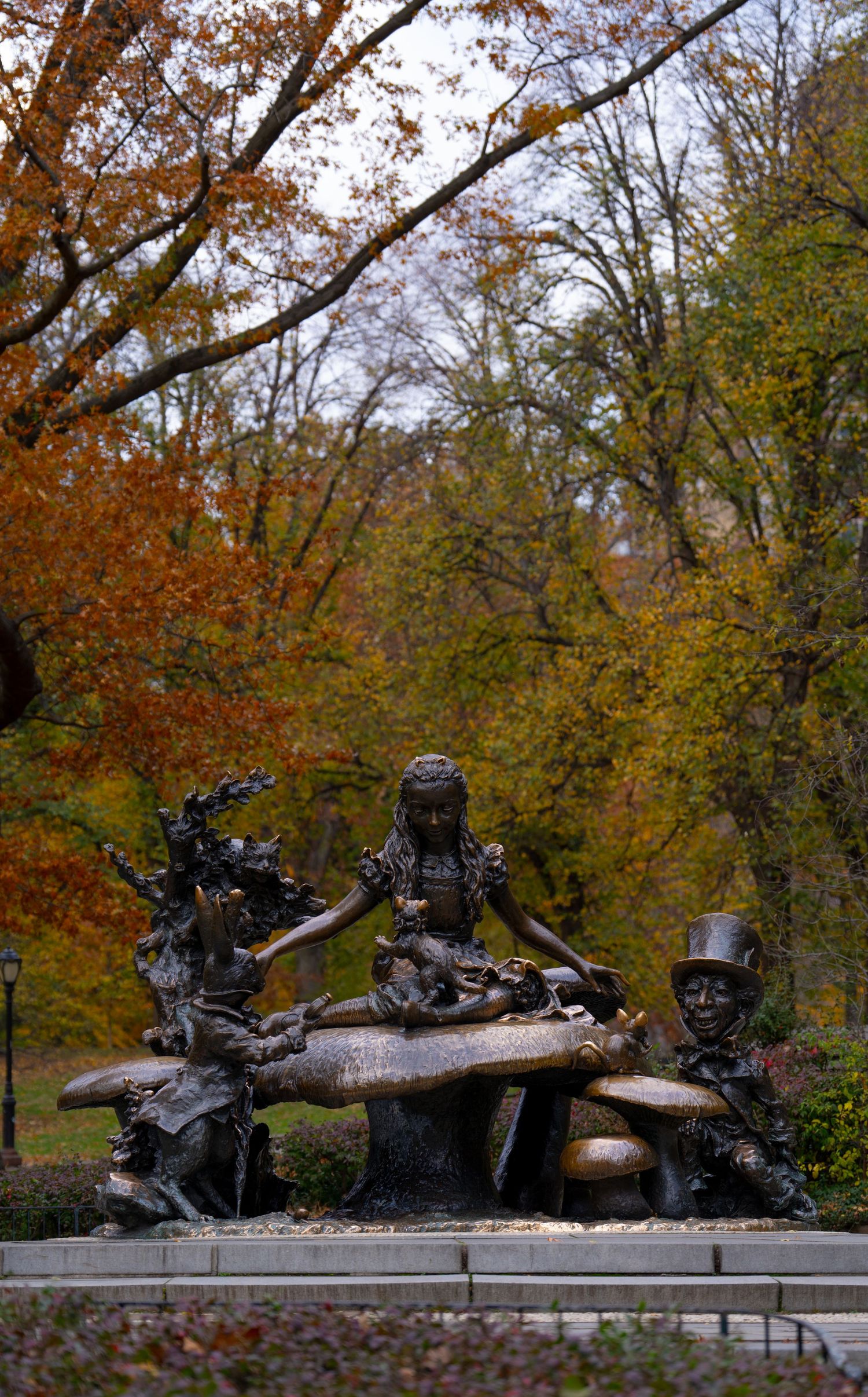 Bronze sculpture installation depicting figures resting on logs in a park surrounded by autumn foliage.