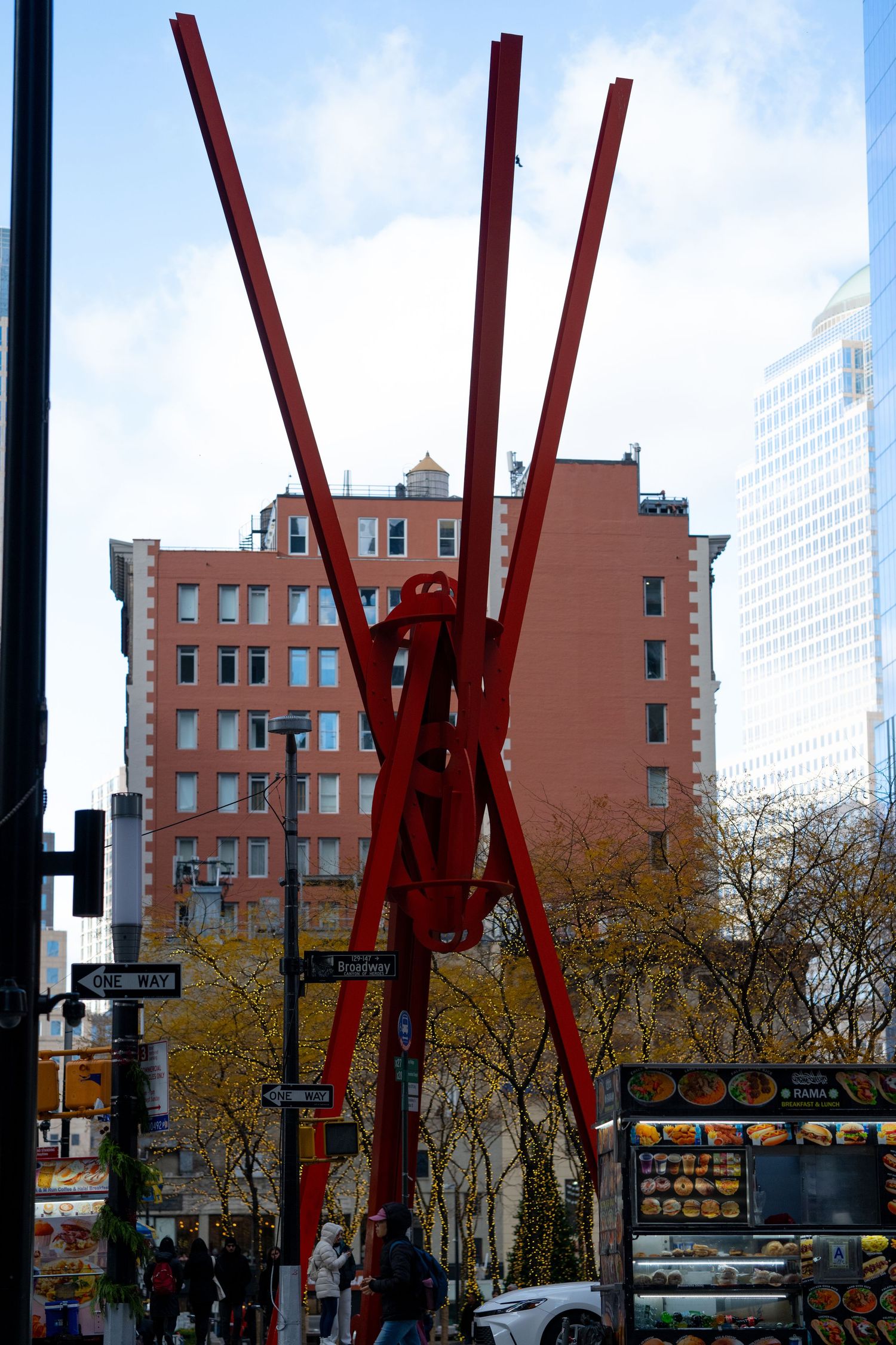A tall red metal sculpture with intersecting beams stands prominently against an urban backdrop with brick buildings.