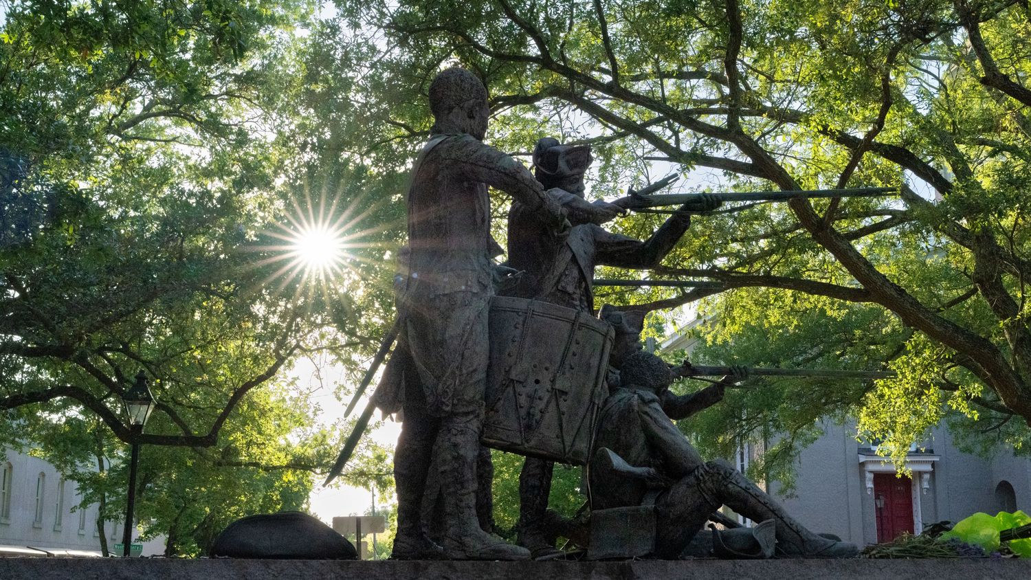 Park statues and monuments under lush green trees with sunlight streaming through branches.