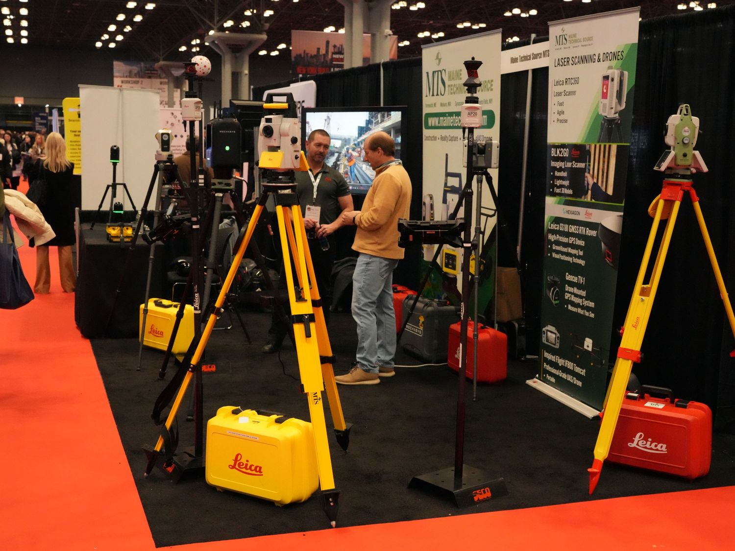 Leica surveying equipment display at a trade show with tripods and yellow cases on a red carpet floor.