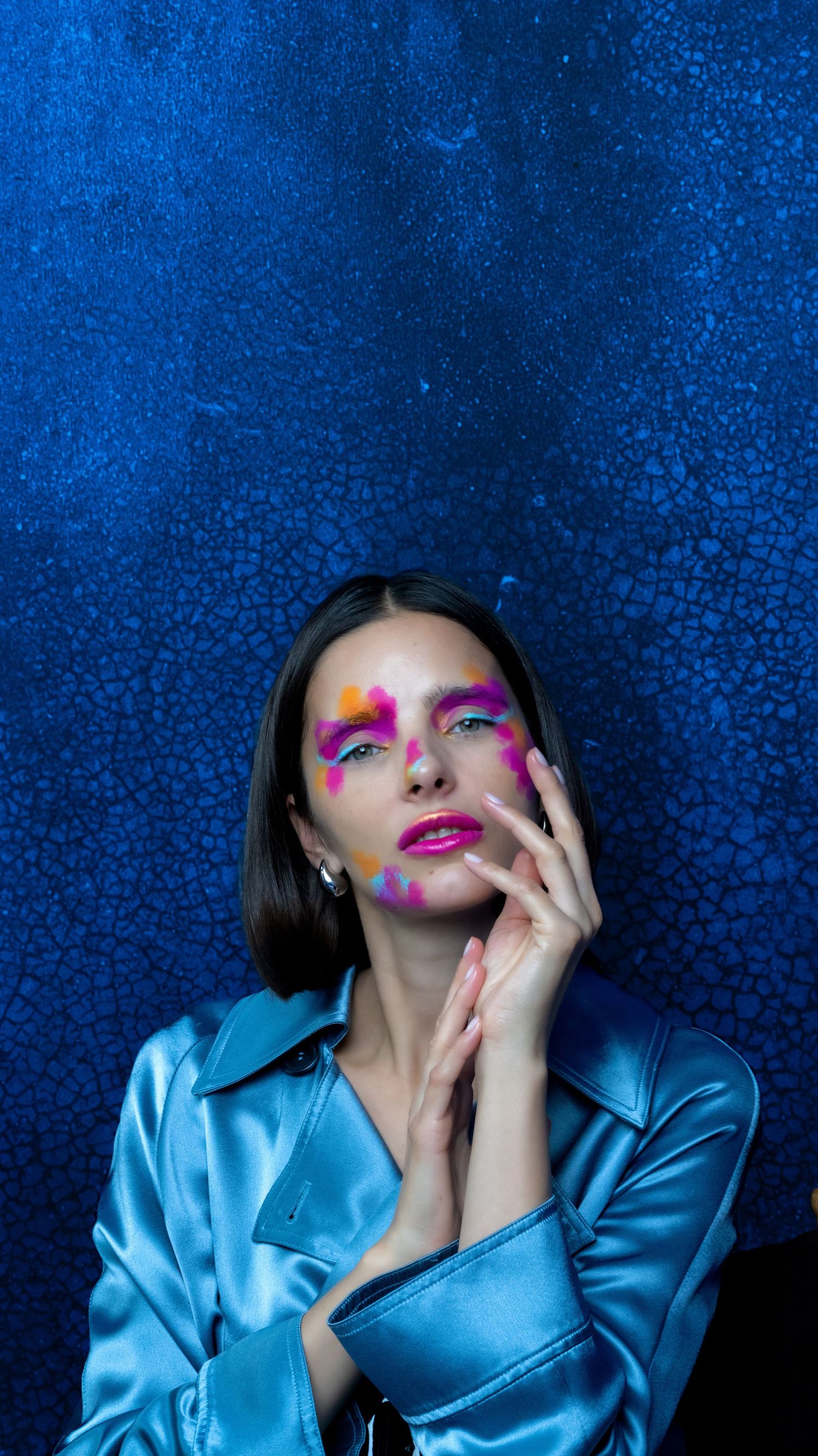 Artistic fashion photo with vibrant pink makeup and blue satin shirt against dark backdrop.