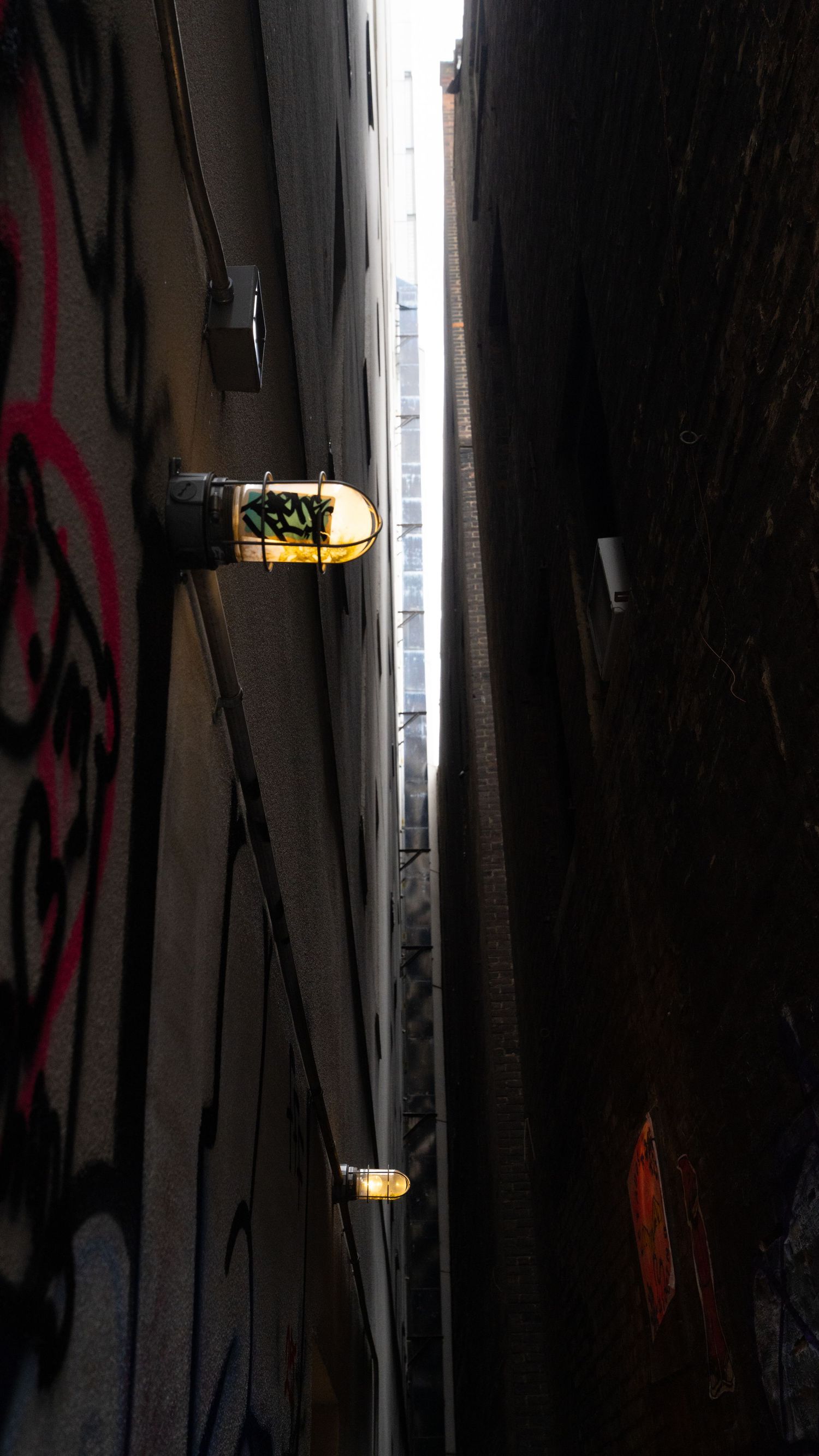 Dark narrow alley between buildings with dim lighting fixtures visible.