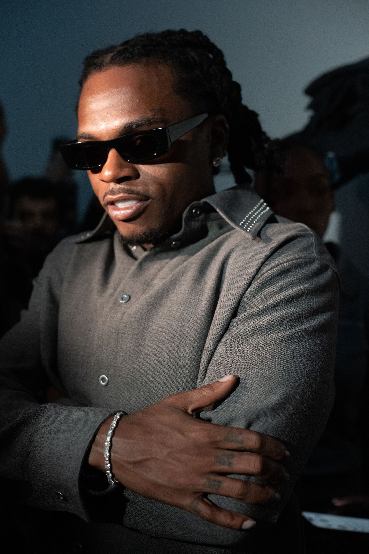 Figure in grey suit jacket crossing arms while wearing sunglasses in dramatic lighting.