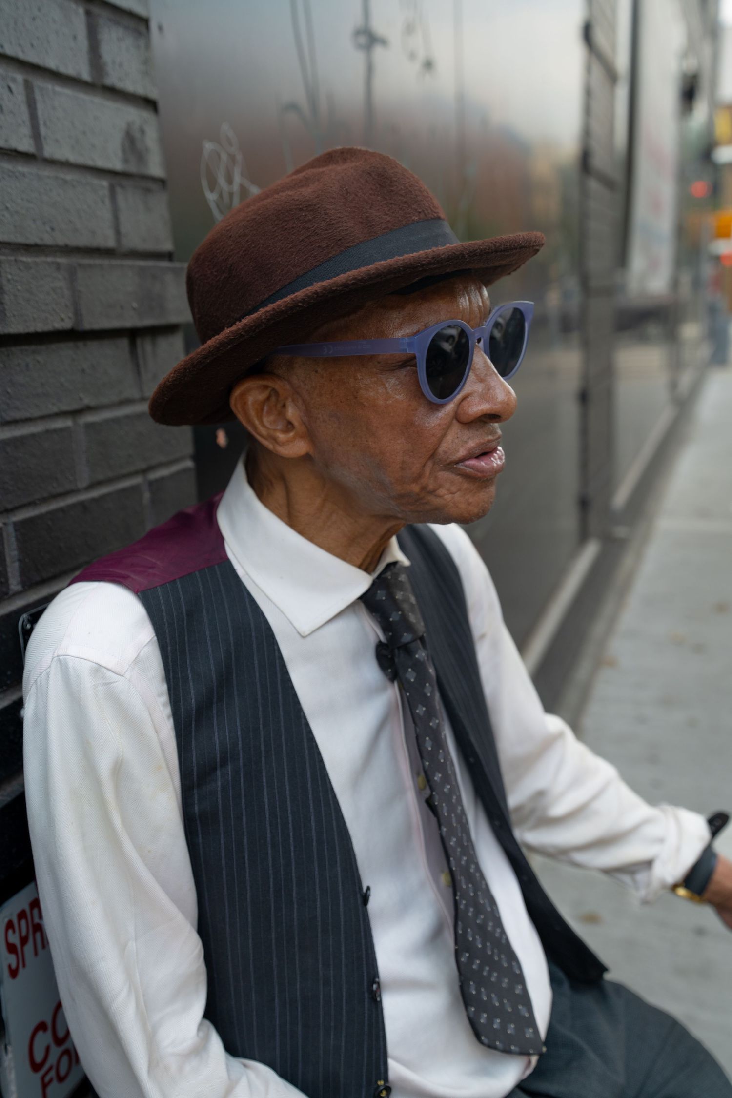 A person in vintage clothing and sunglasses sits against an urban wall in various poses.